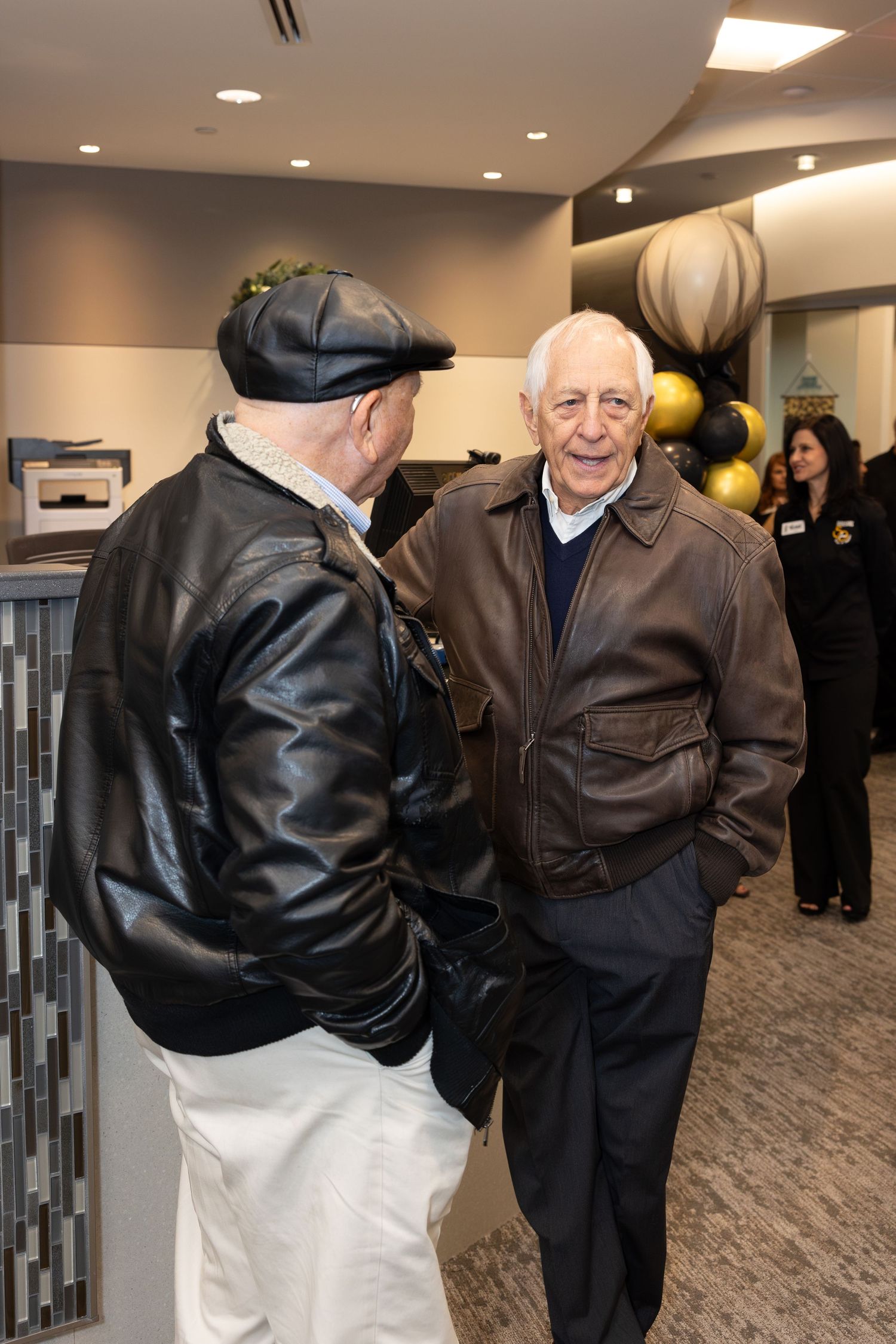 Two men in leather jackets having a conversation in an office hallway at celebration event.