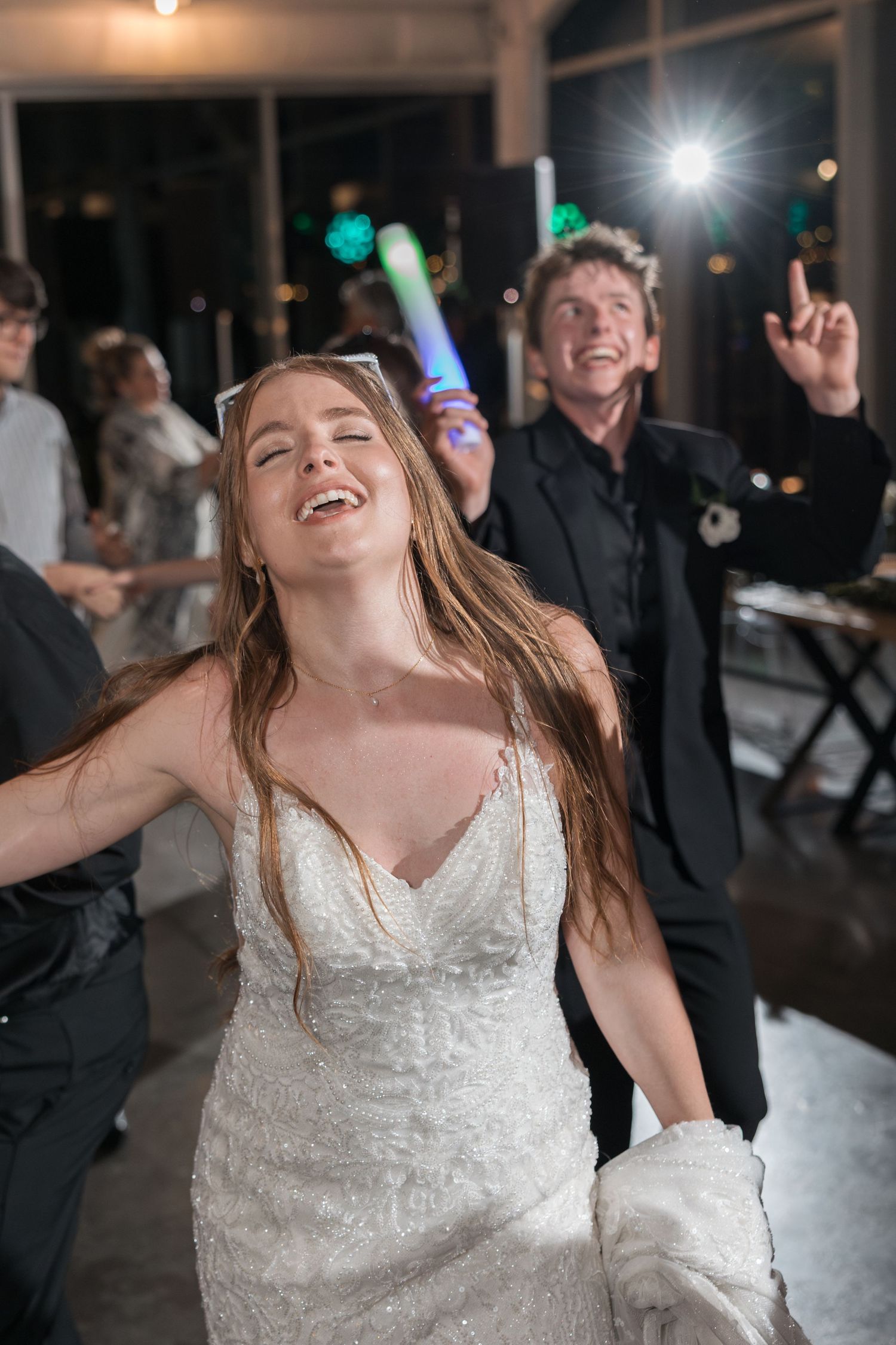 A bride in a white lace dress dances joyfully at her wedding reception with guests celebrating behind her.