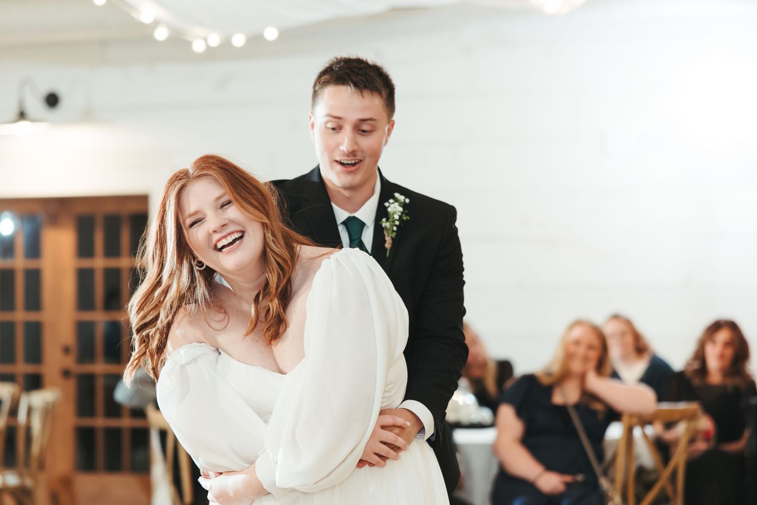A joyful wedding couple laughs while dancing in a bright reception venue.