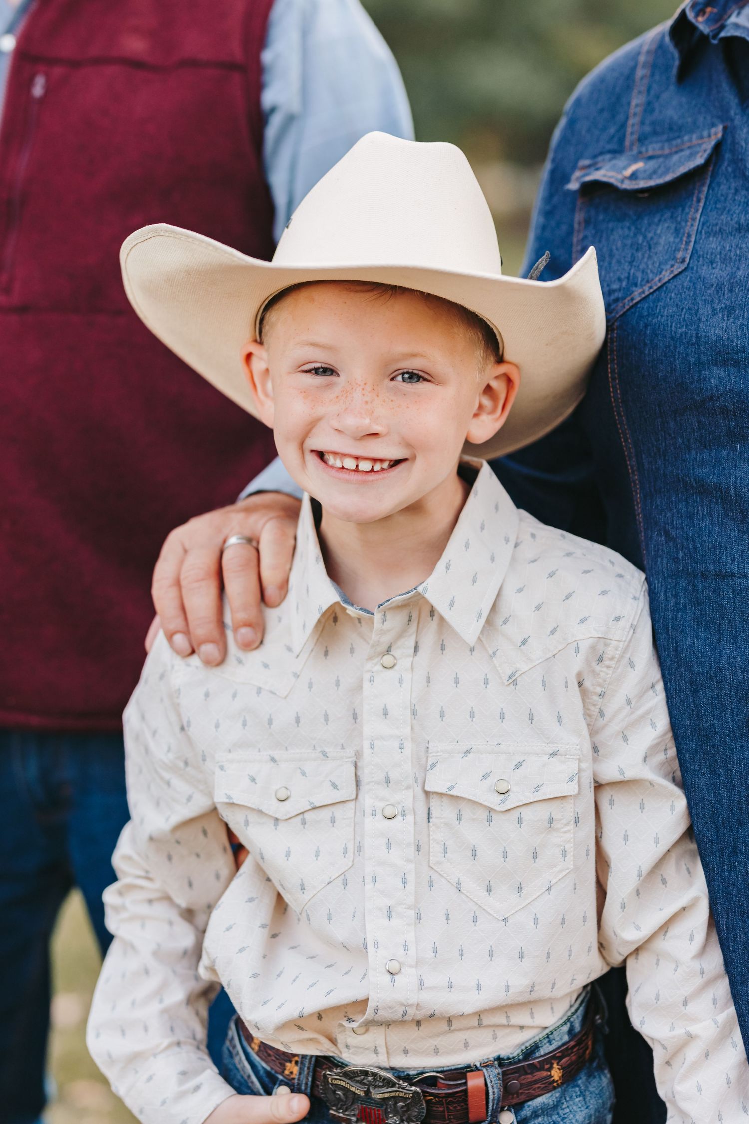 Child in western attire wearing cream colored cowboy hat and button-down shirt smiles at camera.