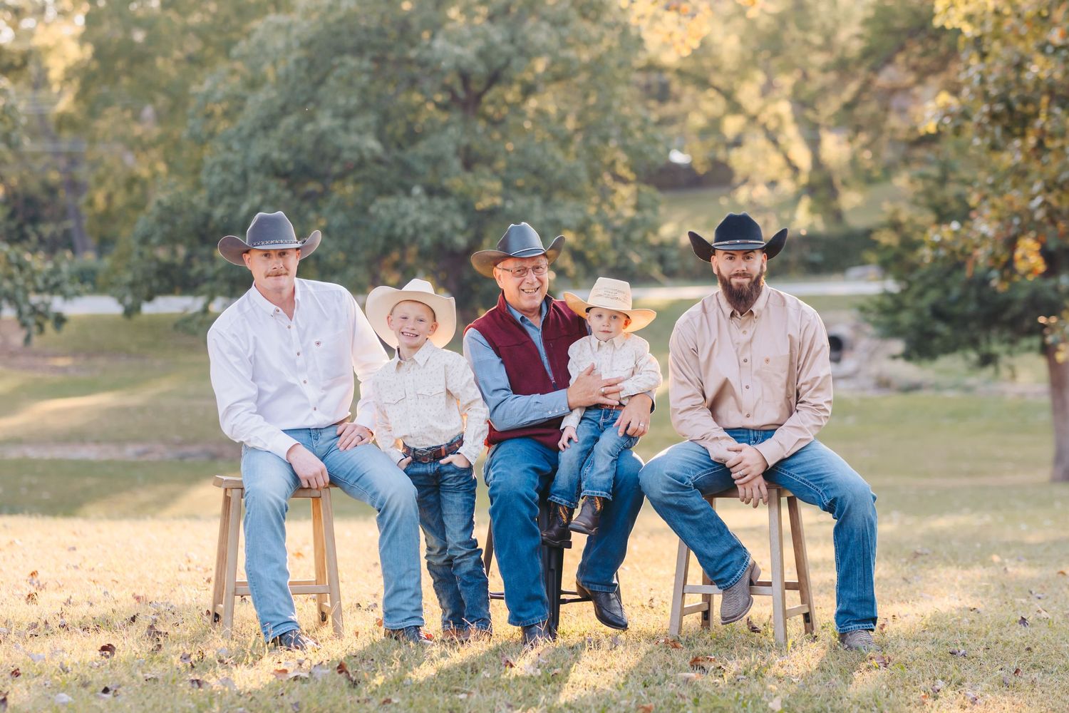A group sits together on wooden stools wearing western attire with cowboy hats in an outdoor autumn setting.