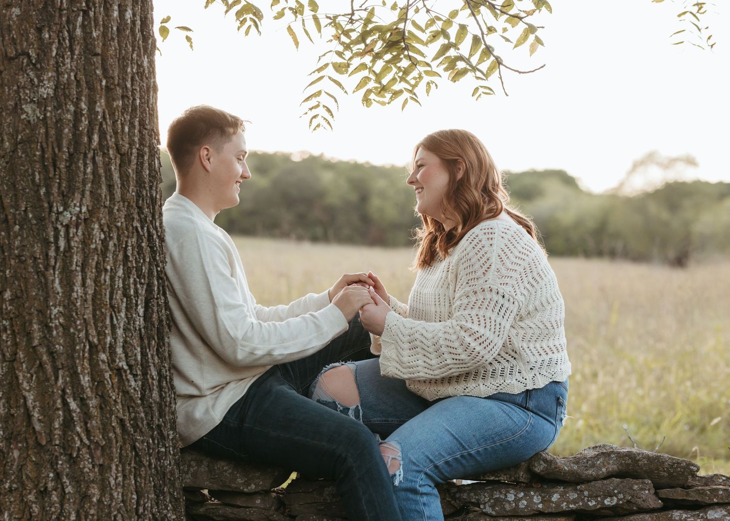 A couple sits together under a tree in a sunny field.