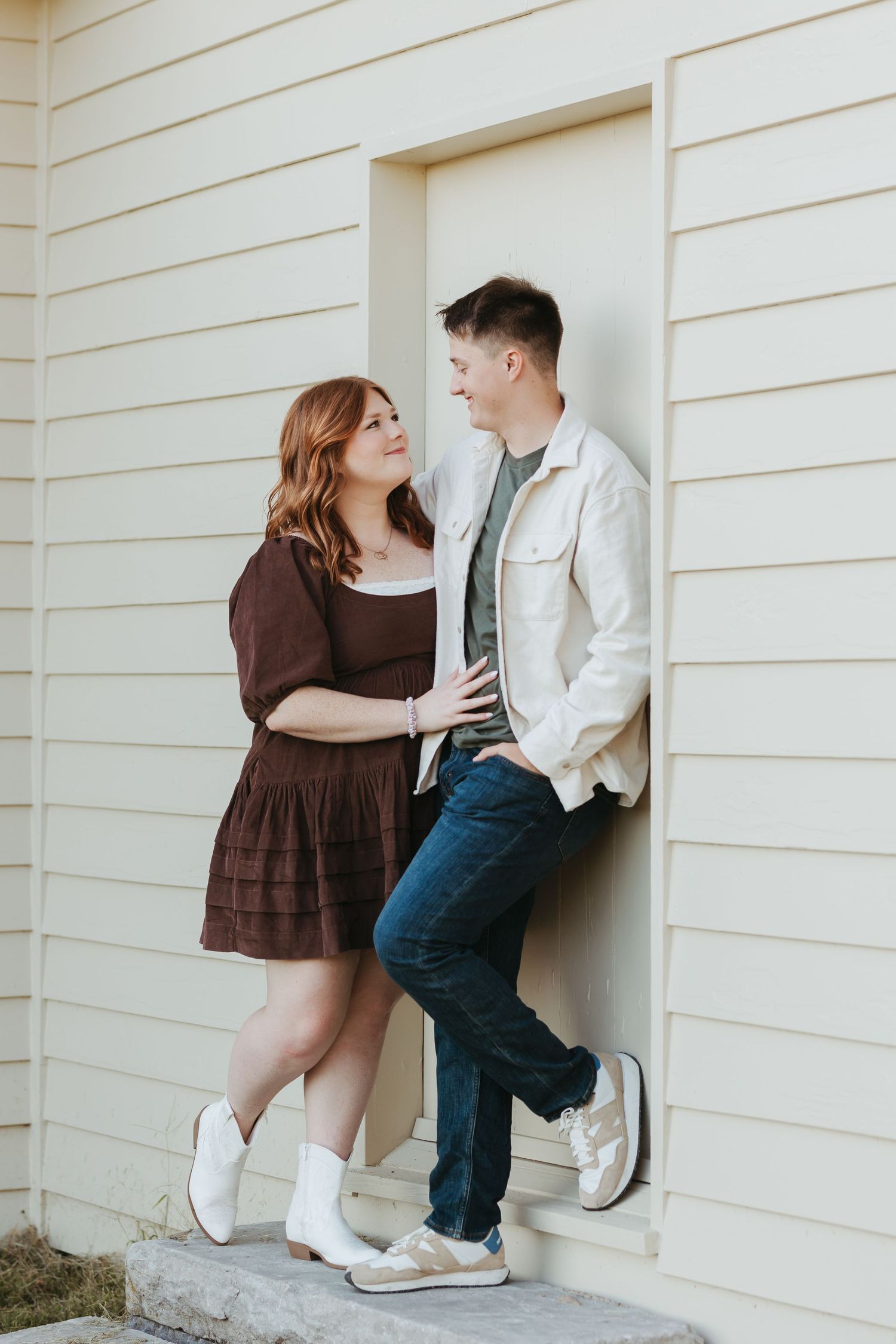 Couple in brown dress and white jacket share intimate moment against house exterior.