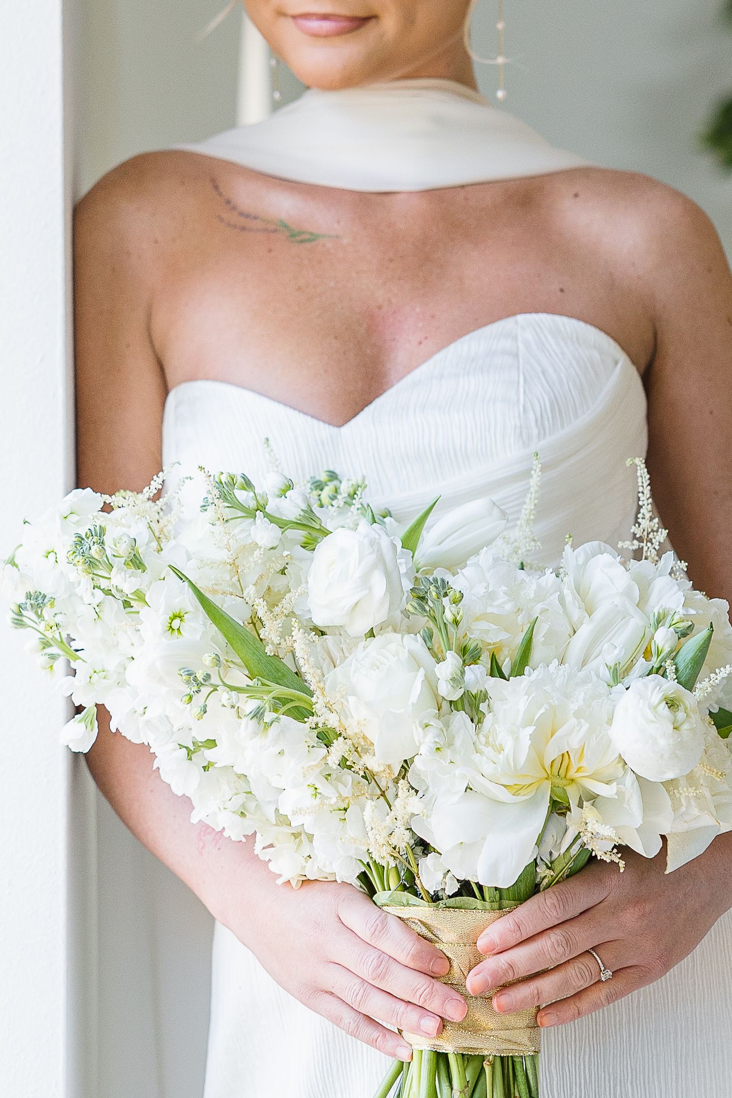 A white bridal bouquet with roses and delicate blooms is held against a strapless white wedding gown.