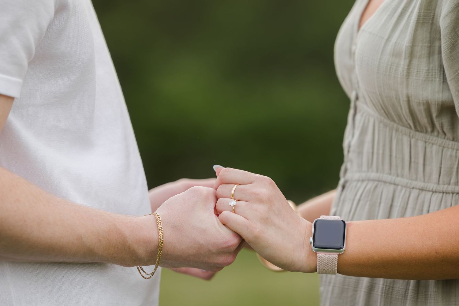 Close-up of two people holding hands outdoors, one wearing white and one wearing a smartwatch and beige dress.