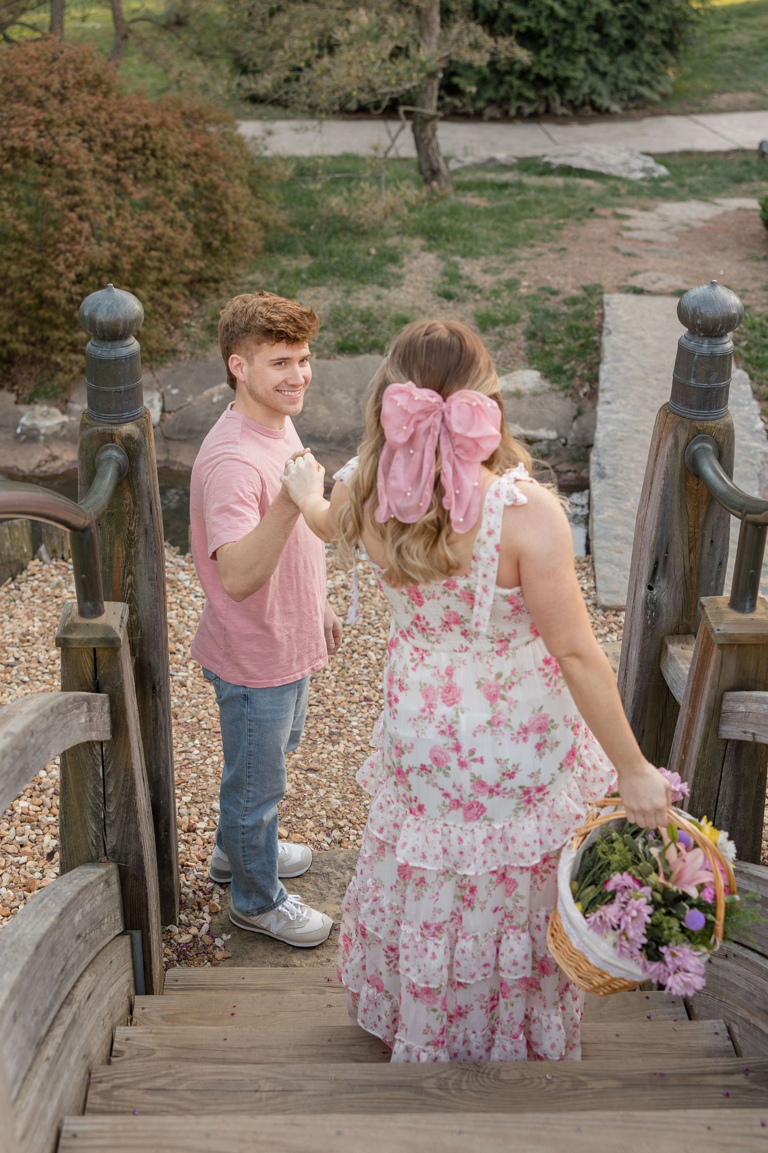 A couple stands on a wooden bridge, with one holding a basket of flowers while wearing a floral dress on a sunny day.
