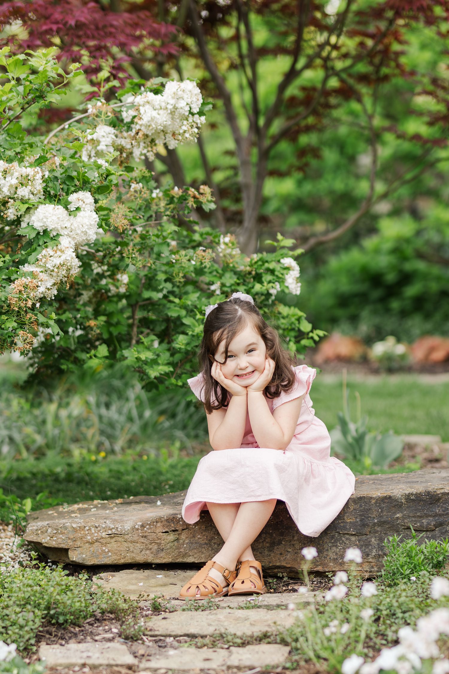 A child wearing a pink dress sits thoughtfully on stone steps in a garden with blooming white flowers.