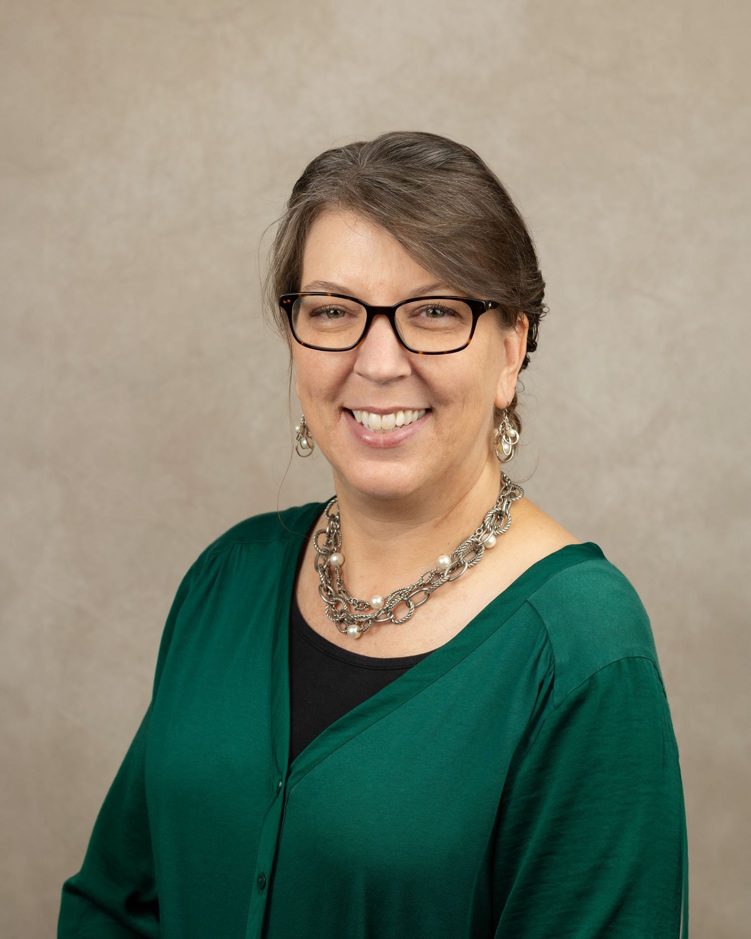 Professional headshot of a person in a green cardigan and statement necklace, wearing glasses and smiling warmly.