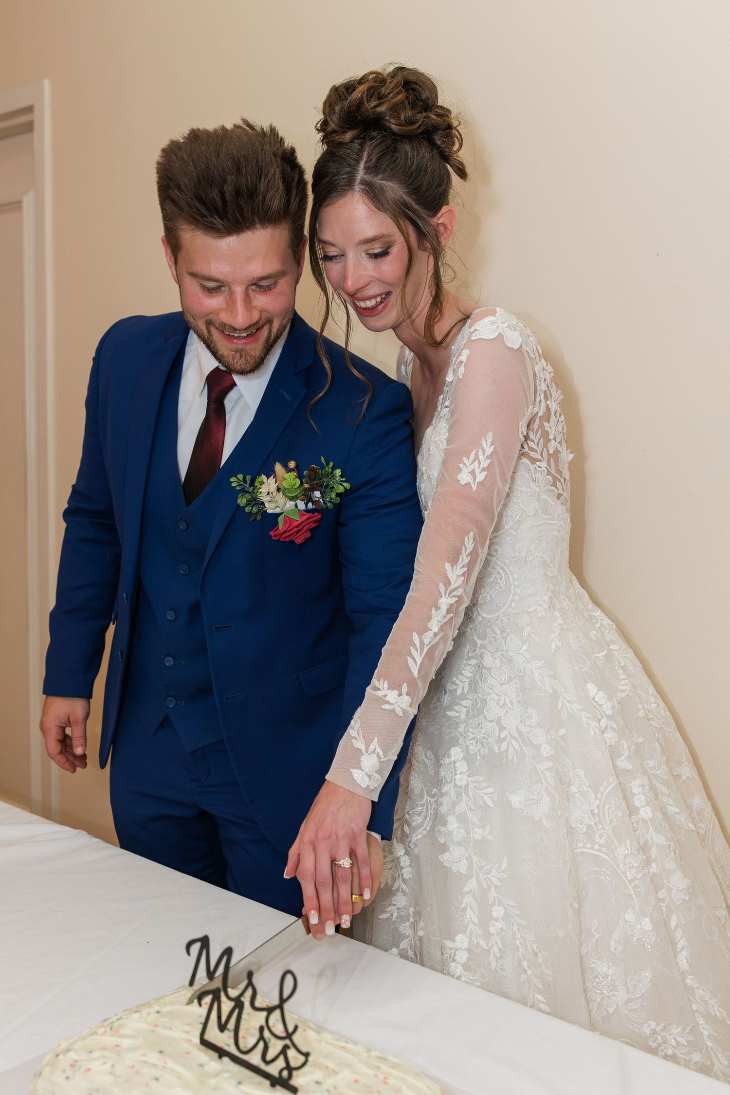 Smiling bride and groom in blue suit cut their wedding cake together at their Mr. and Mrs. reception.