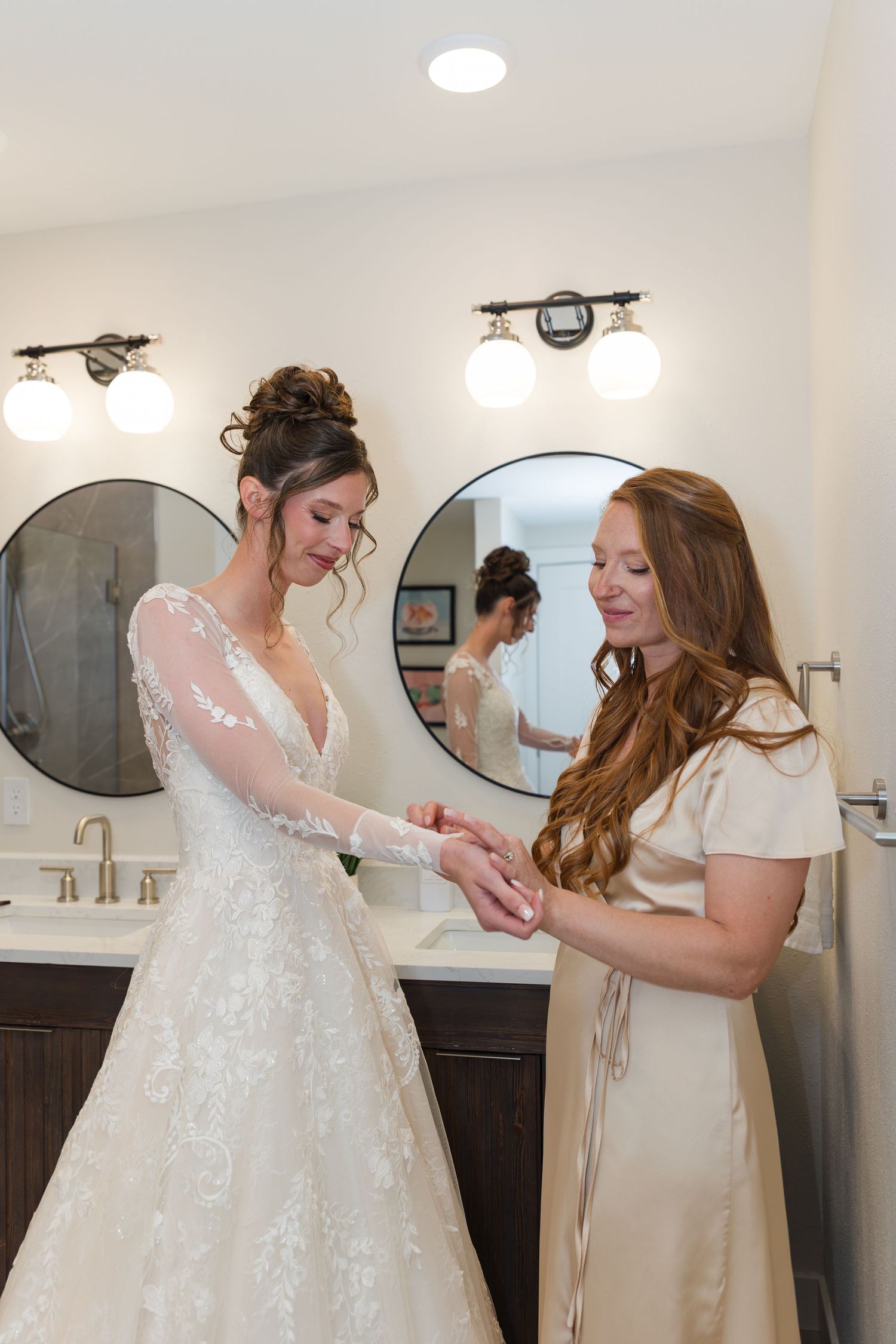 Bride in lace wedding gown gets assisted by redhead bridesmaid in champagne dress in elegant bathroom.