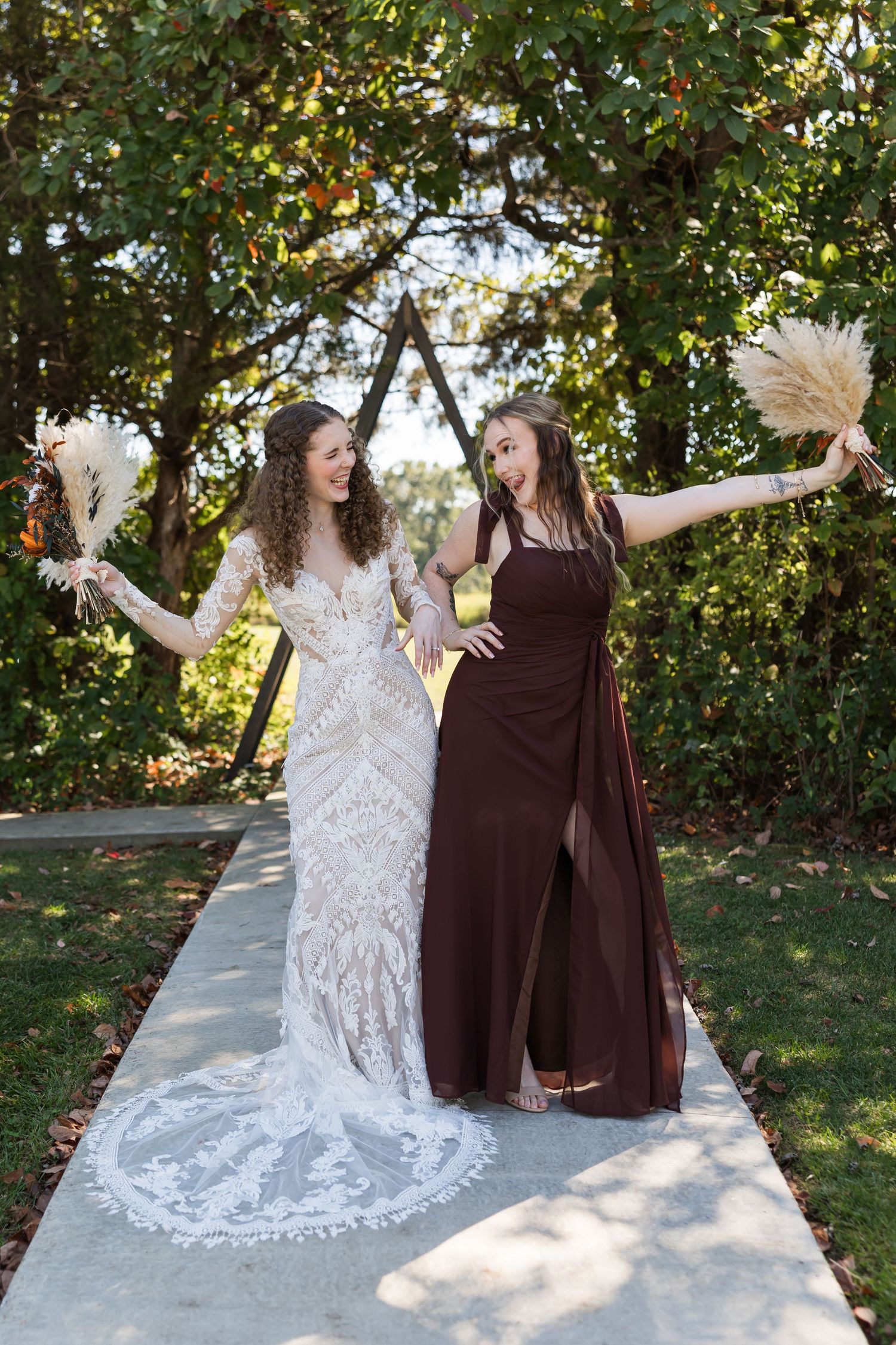 Bride in lace gown and bridesmaid in burgundy dress pose joyfully on garden pathway holding pampas grass bouquets.