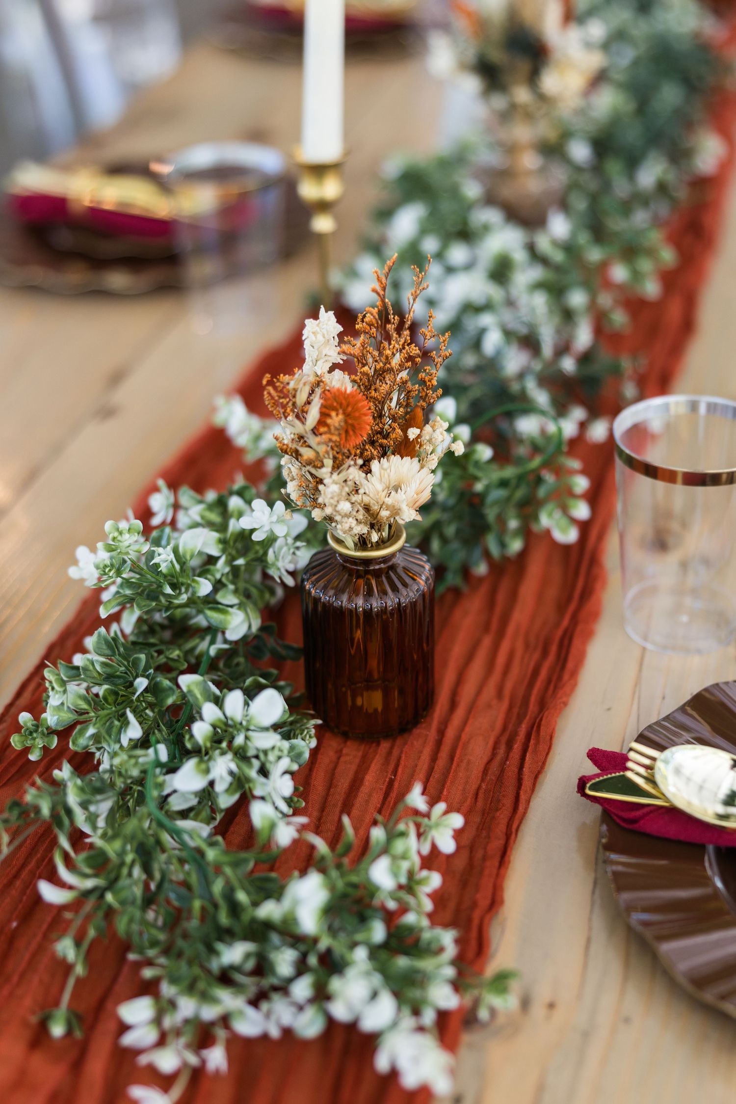 Rustic wedding table with terracotta runner, greenery garland, dried flowers in amber vase, and gold candlesticks on wood table.