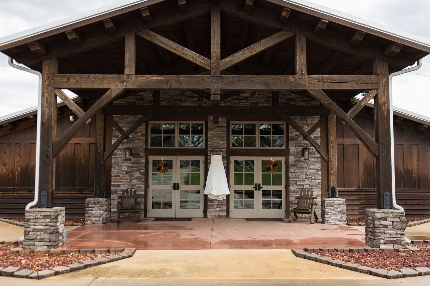 Rustic wooden wedding venue exterior with stone accents, vaulted timber frame entrance, and bridal gown hanging on glass doors.