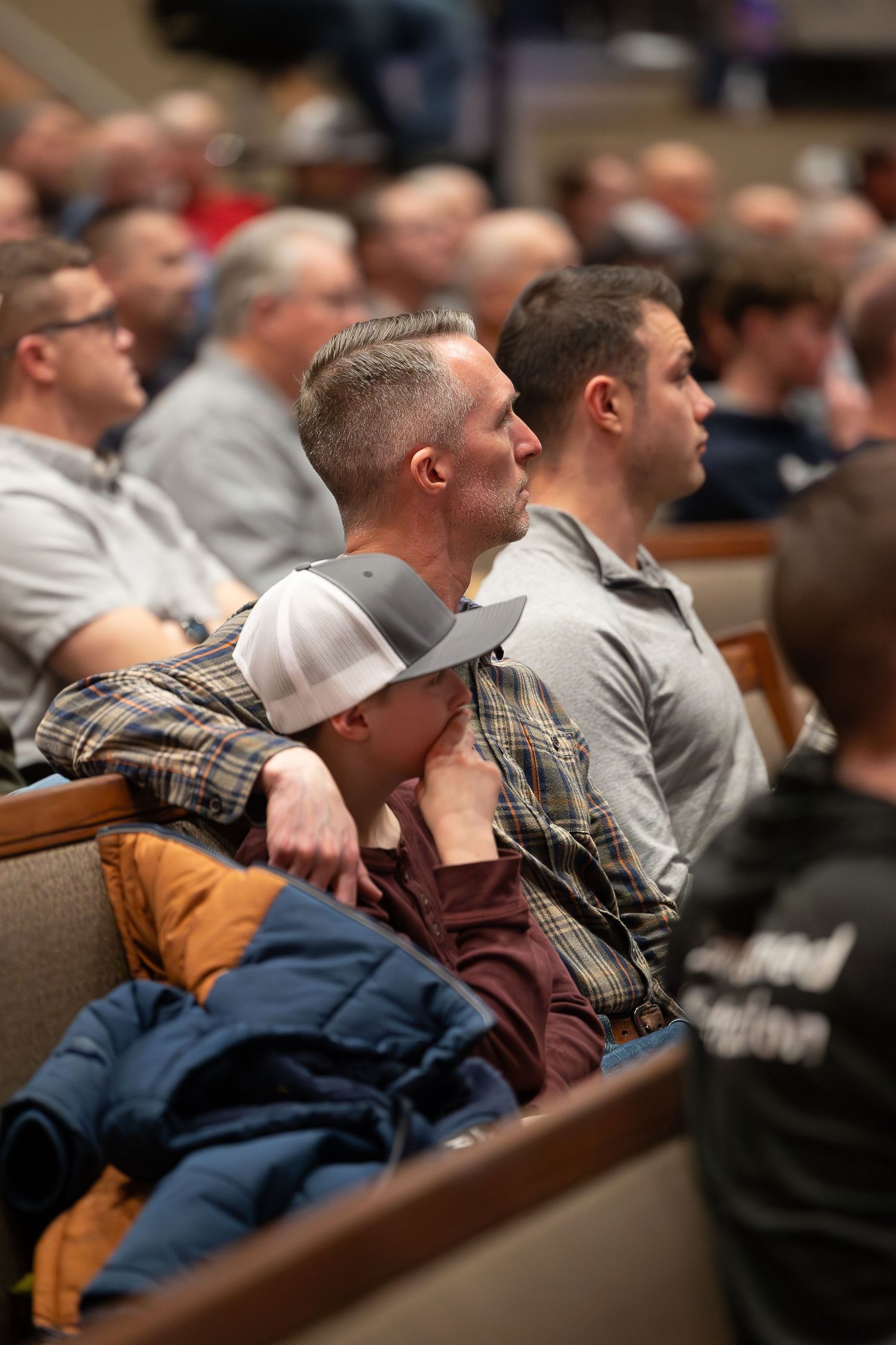 People sit attentively in rows of church or auditorium pews during what appears to be a gathering or service.