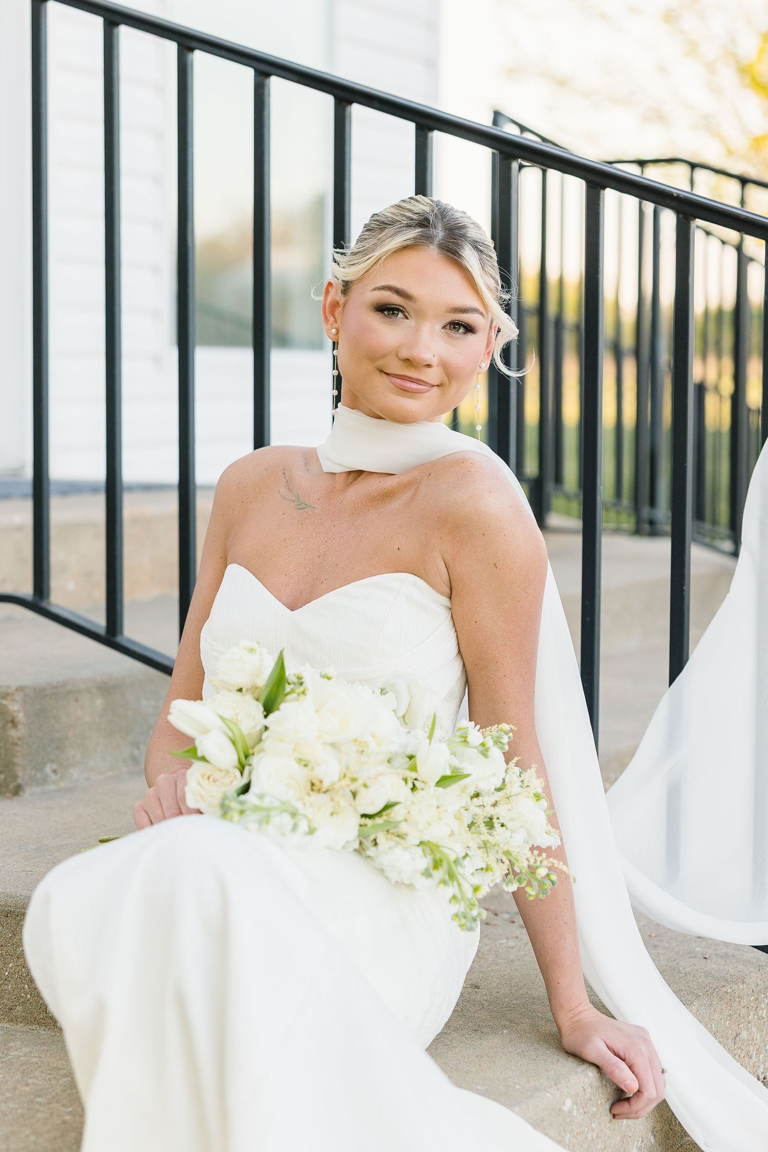 A bride in a strapless white gown holds a luxurious white bouquet while sitting on outdoor steps near a railing.