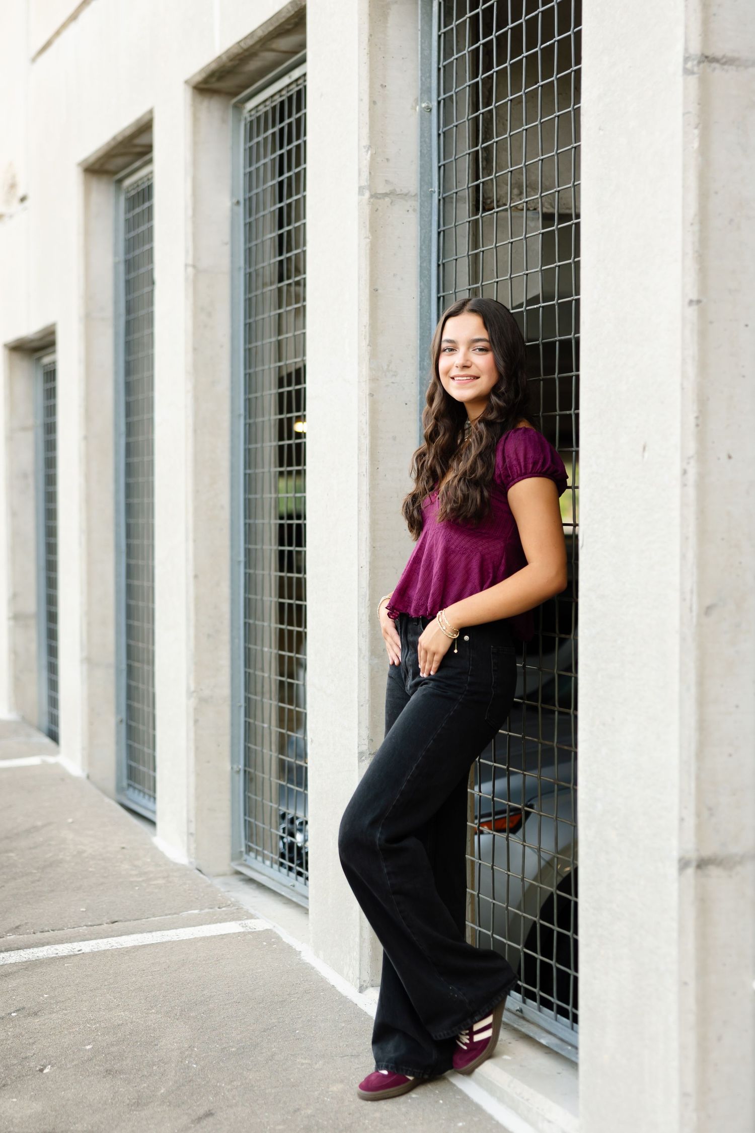 A person in a purple top and black pants poses casually against an industrial building with wire-covered windows.