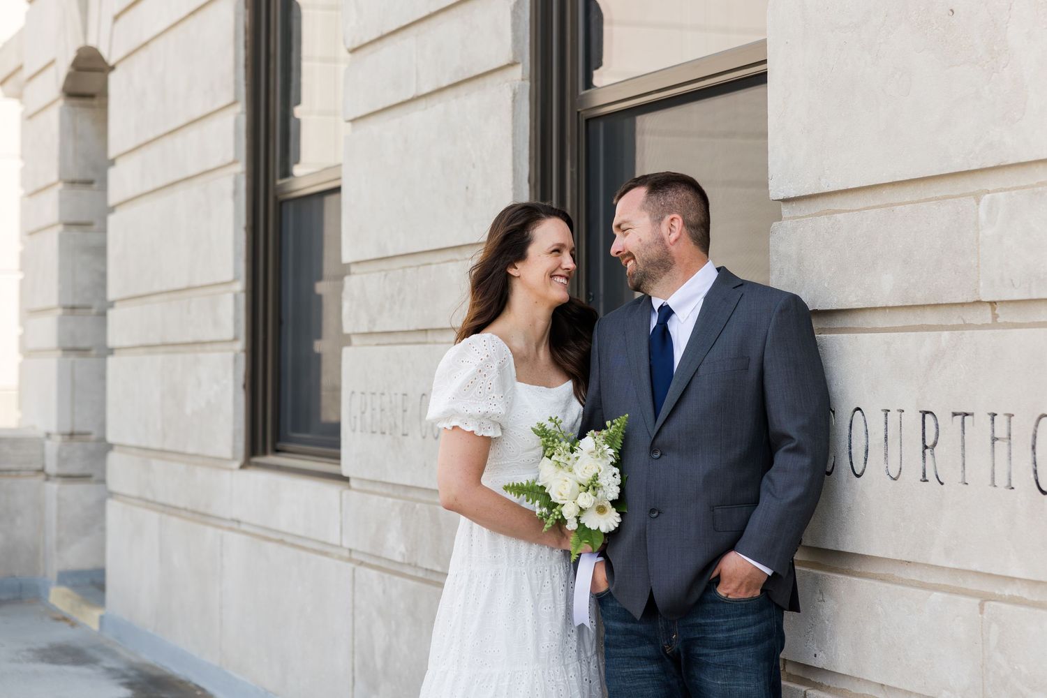A newlywed couple shares a tender moment outside a courthouse building on their wedding day.
