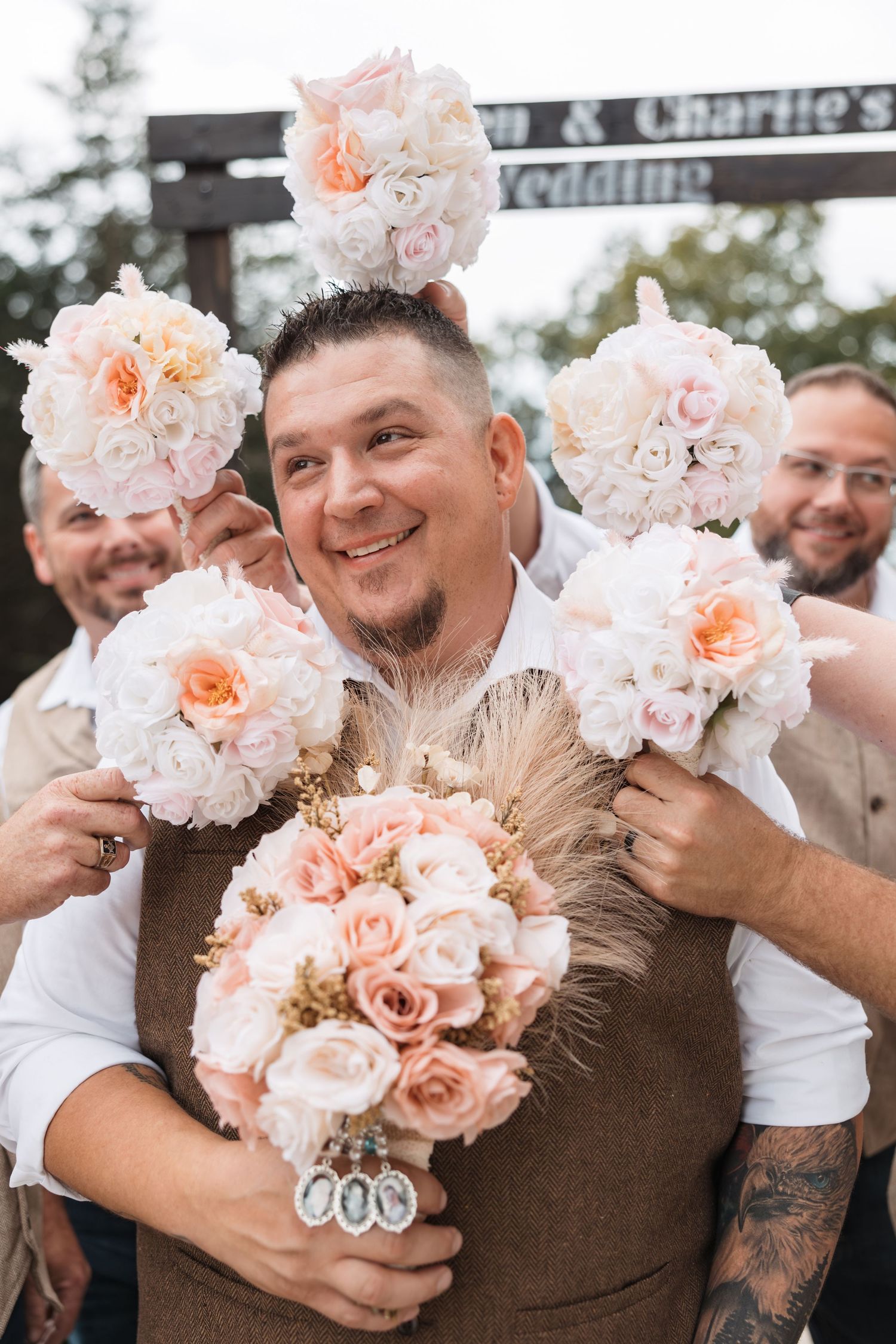Groom smiling surrounded by groomsmen holding pink and white rose bouquets at an outdoor wedding ceremony.