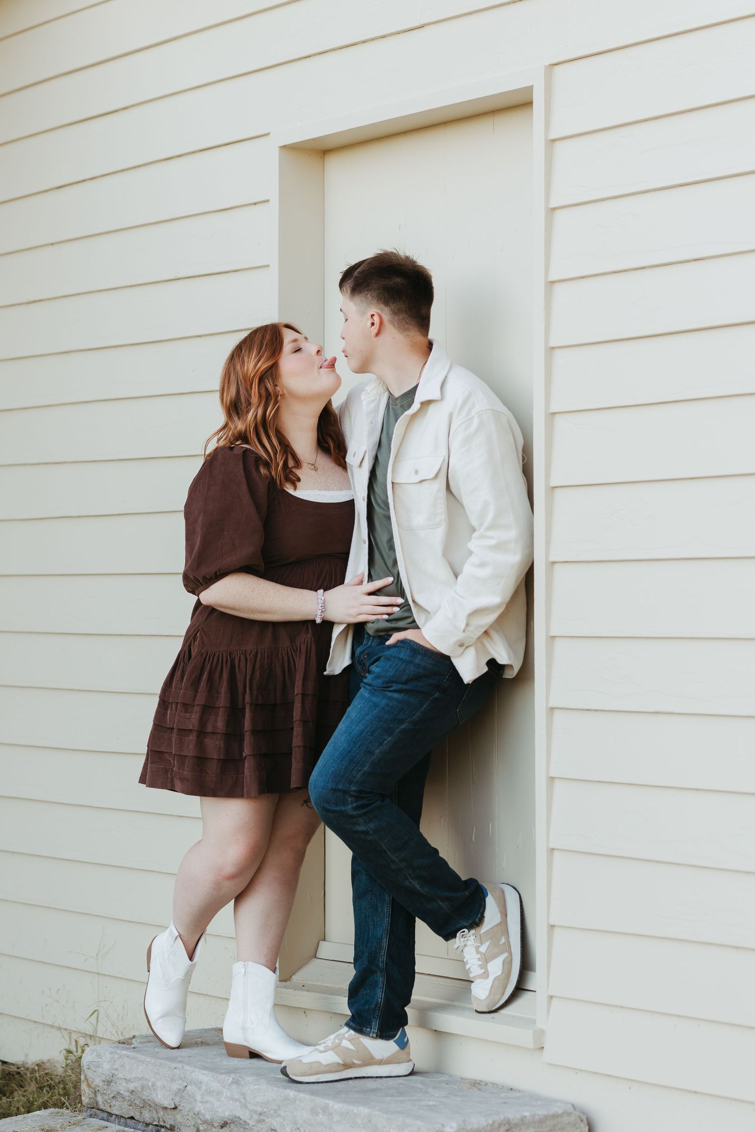 Romantic couple in brown dress and white jacket pose against white house exterior.
