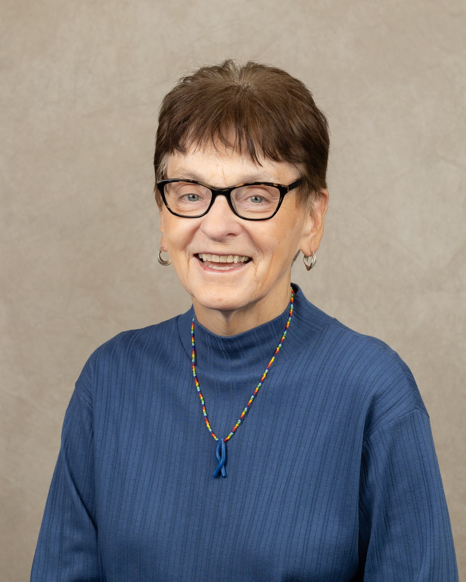 Professional portrait of a person in blue turtleneck wearing glasses and beaded necklace.