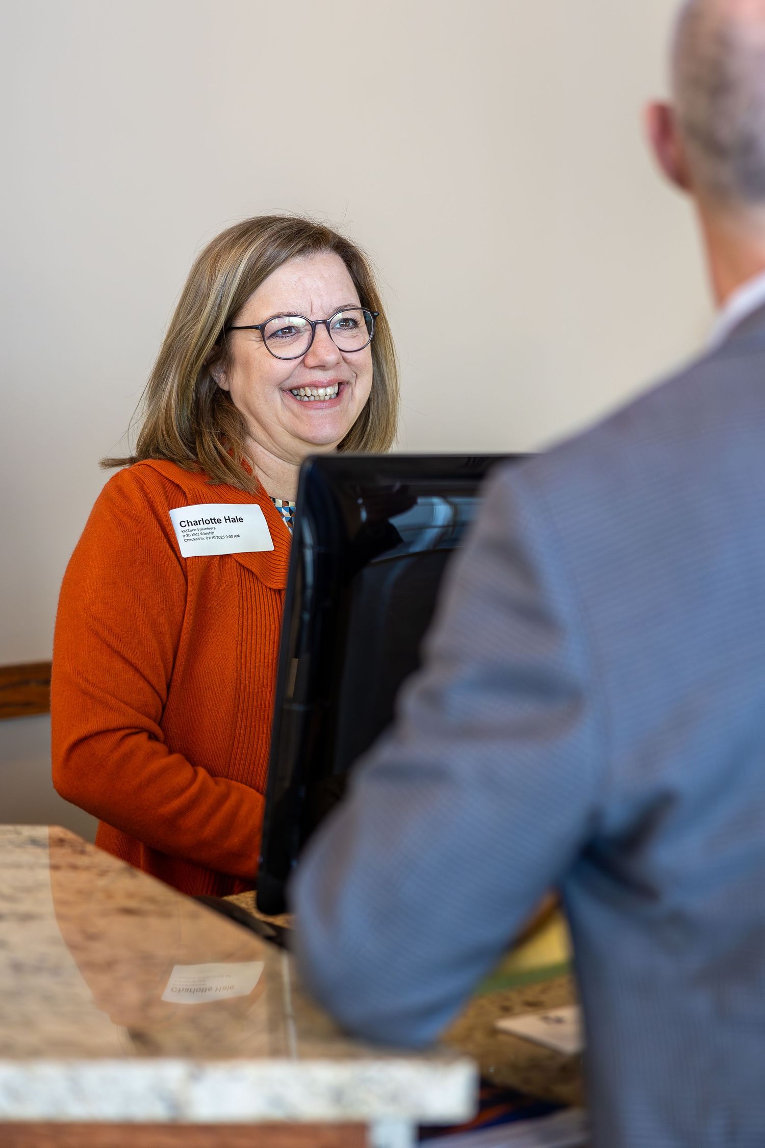 A warm greeting exchanged at a reception desk showing someone in an orange sweater and glasses.