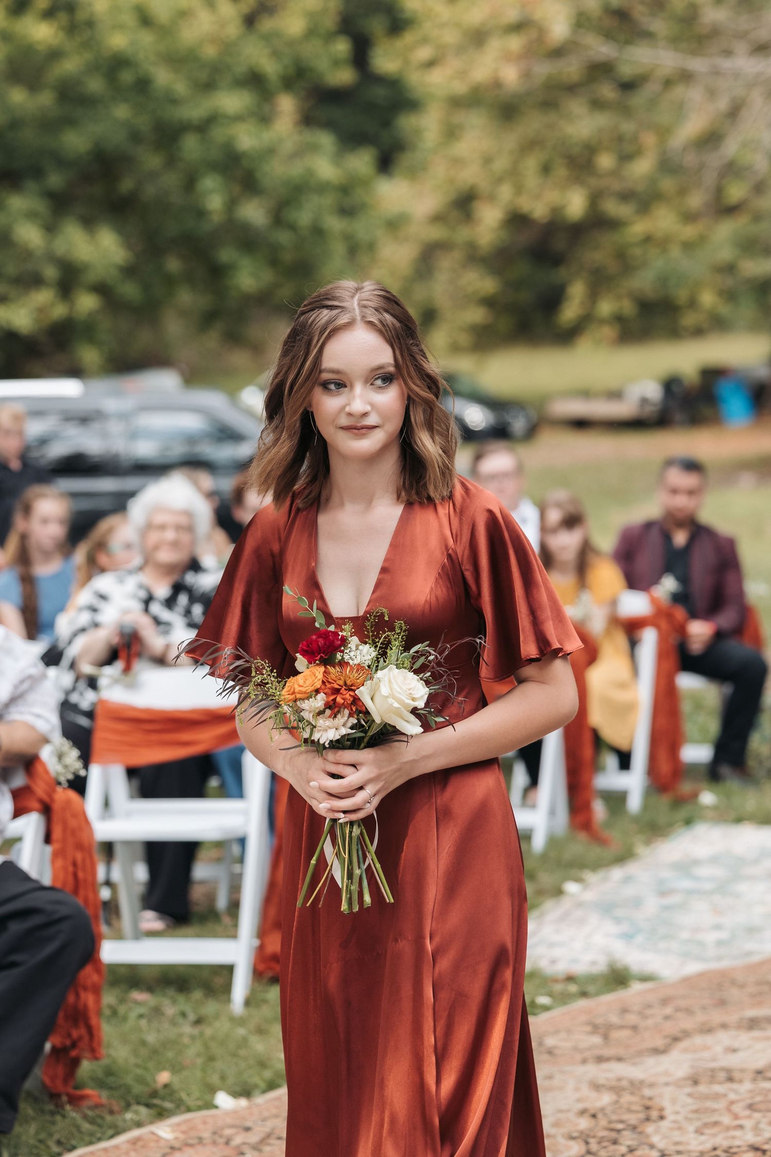 Bridesmaid in rust-colored flutter sleeve dress carries autumn-themed bouquet down outdoor aisle.