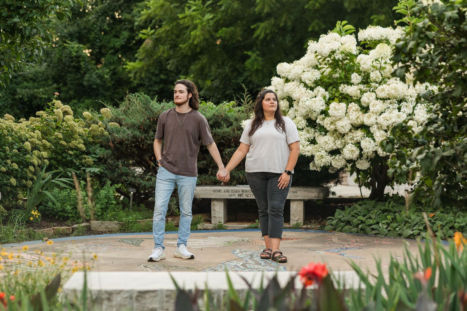 Couple holding hands stands on garden path with blooming white hydrangea bushes in background.