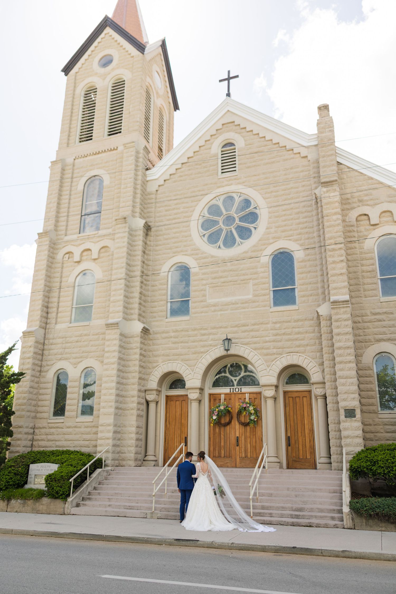 Bride and groom stand before historic stone church with rose window and wooden doors on wedding day.