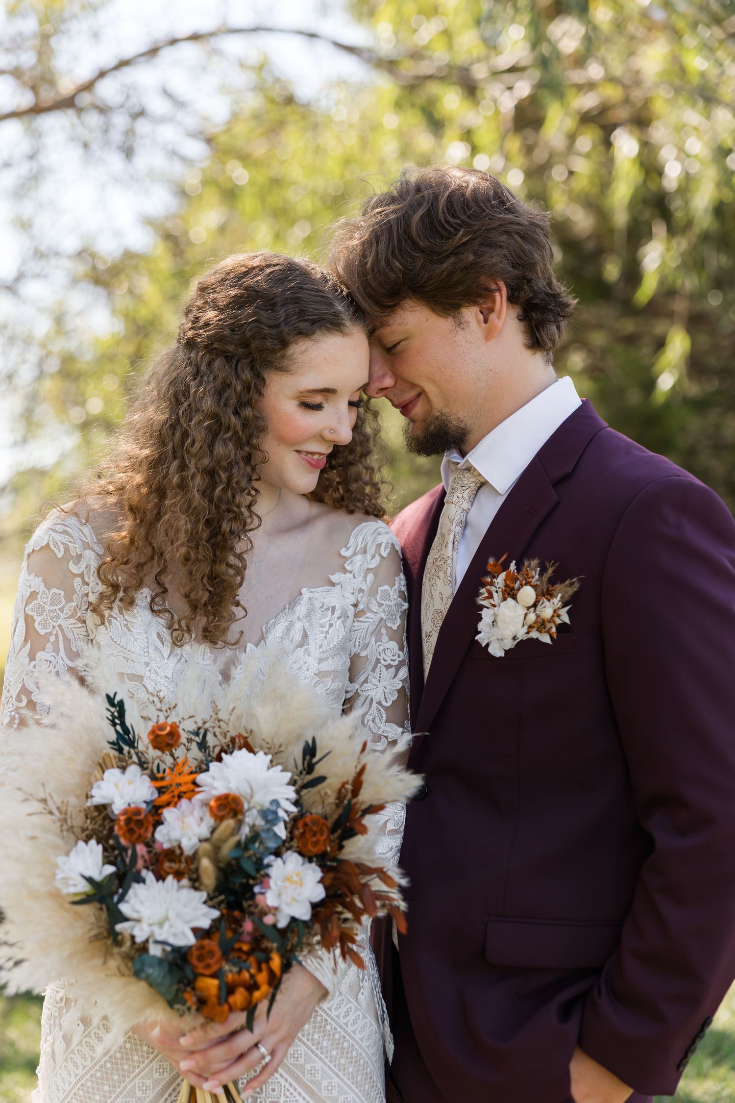 Bride and groom in elegant wedding attire share a romantic moment outdoors, she holds a boho bouquet with pampas grass.