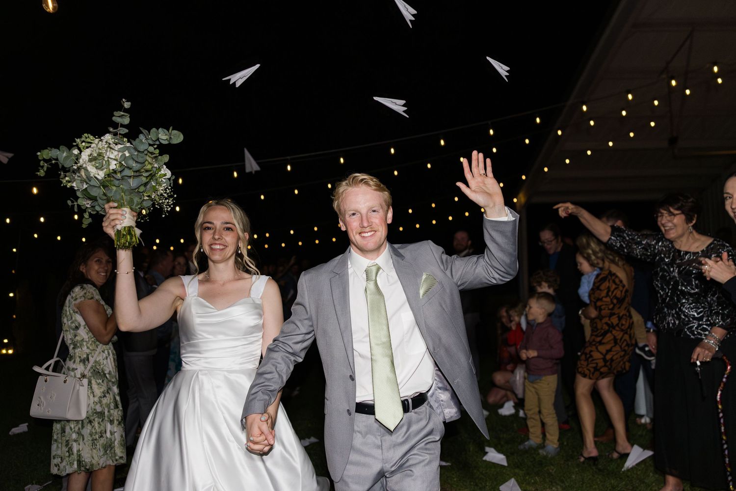 Newlywed couple exits wedding reception as guests toss paper airplanes under string lights at night.