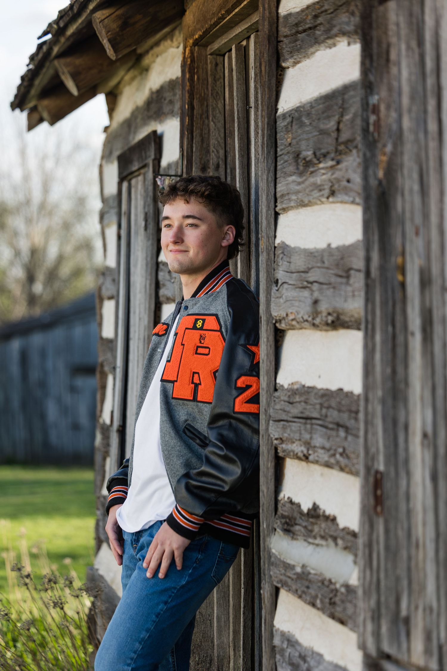 Teen boy in varsity jacket leaning against rustic log cabin wall, smiling confidently in jeans outdoors.