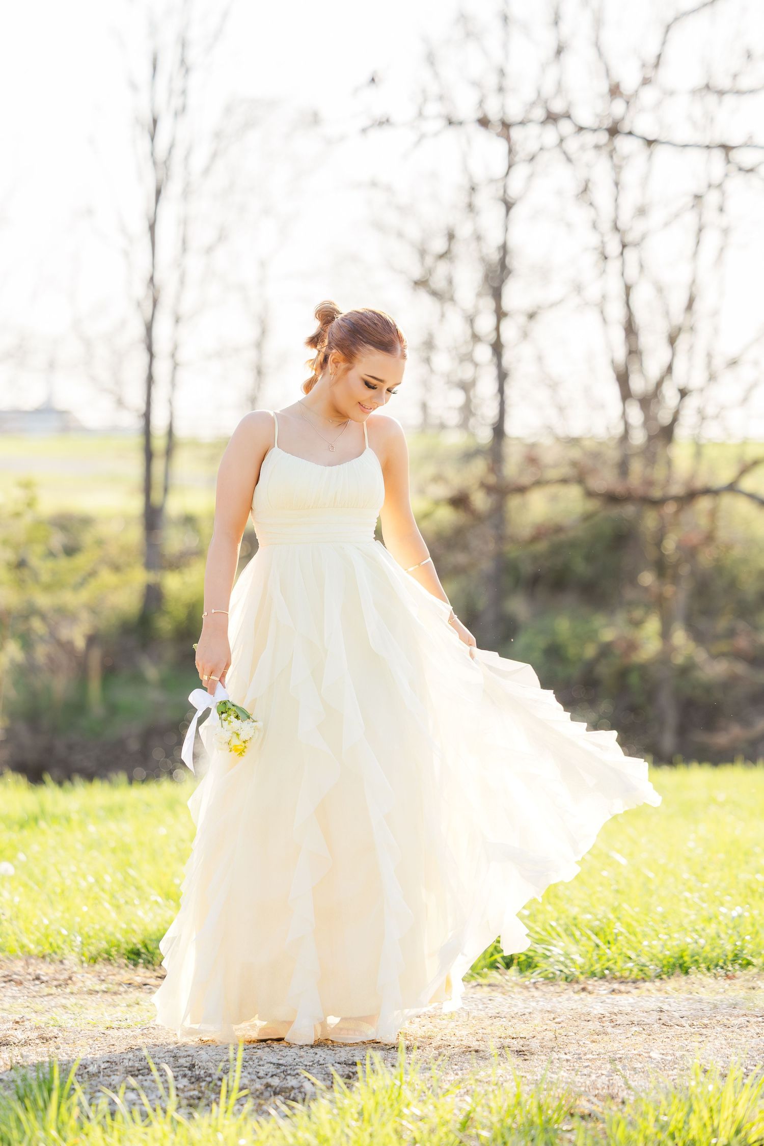 A person in a flowing white wedding dress stands in a sunny meadow with bare trees in the background.