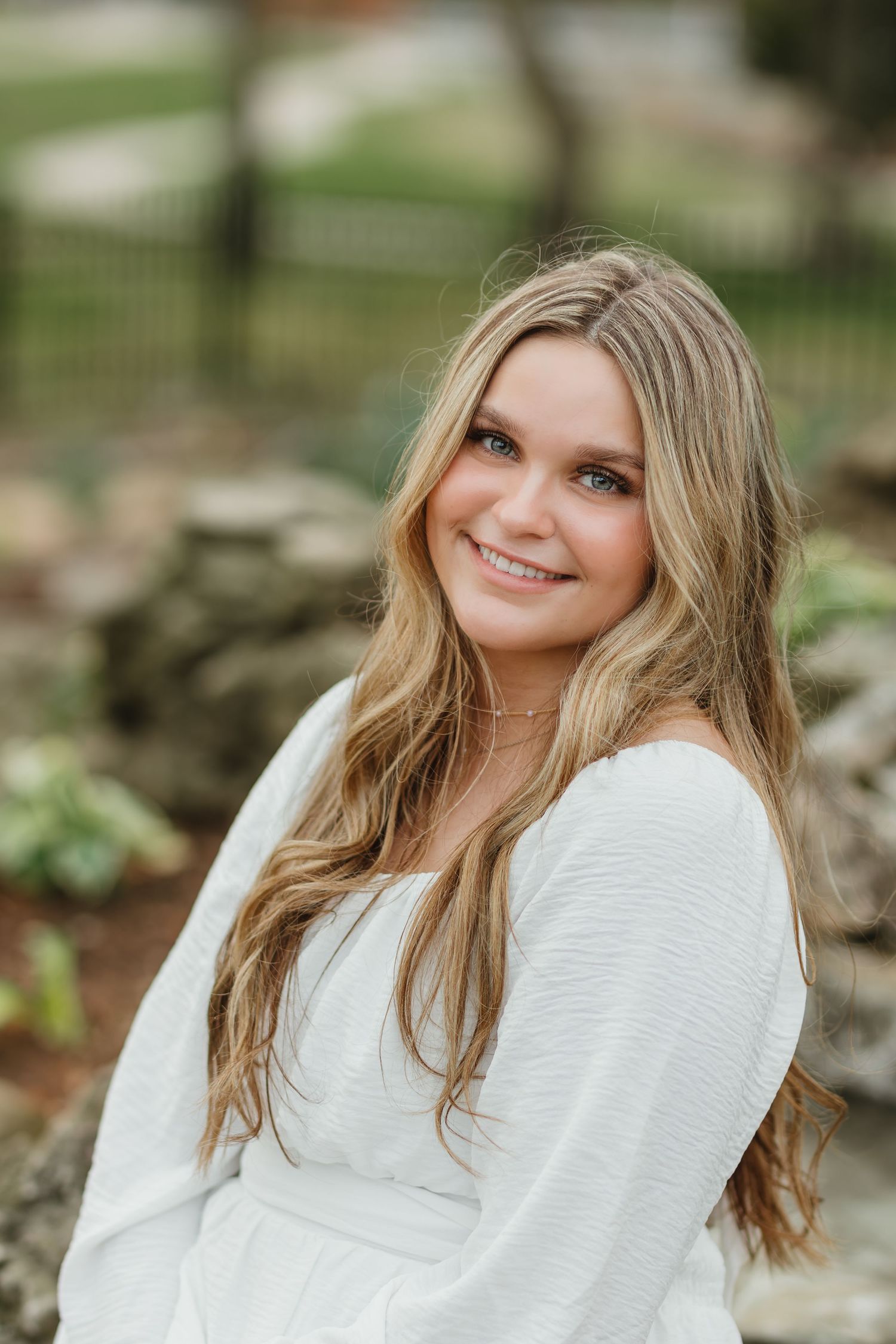 Young woman in white sweater smiling outdoors with long blonde hair against natural garden background.