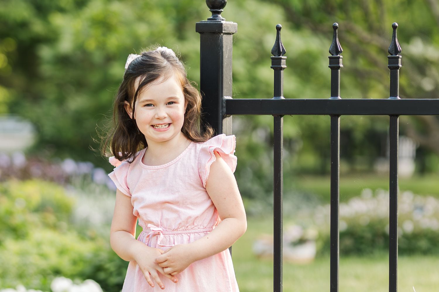 A young girl in a pink ruffled dress smiles while standing next to a black iron gate in a garden setting.