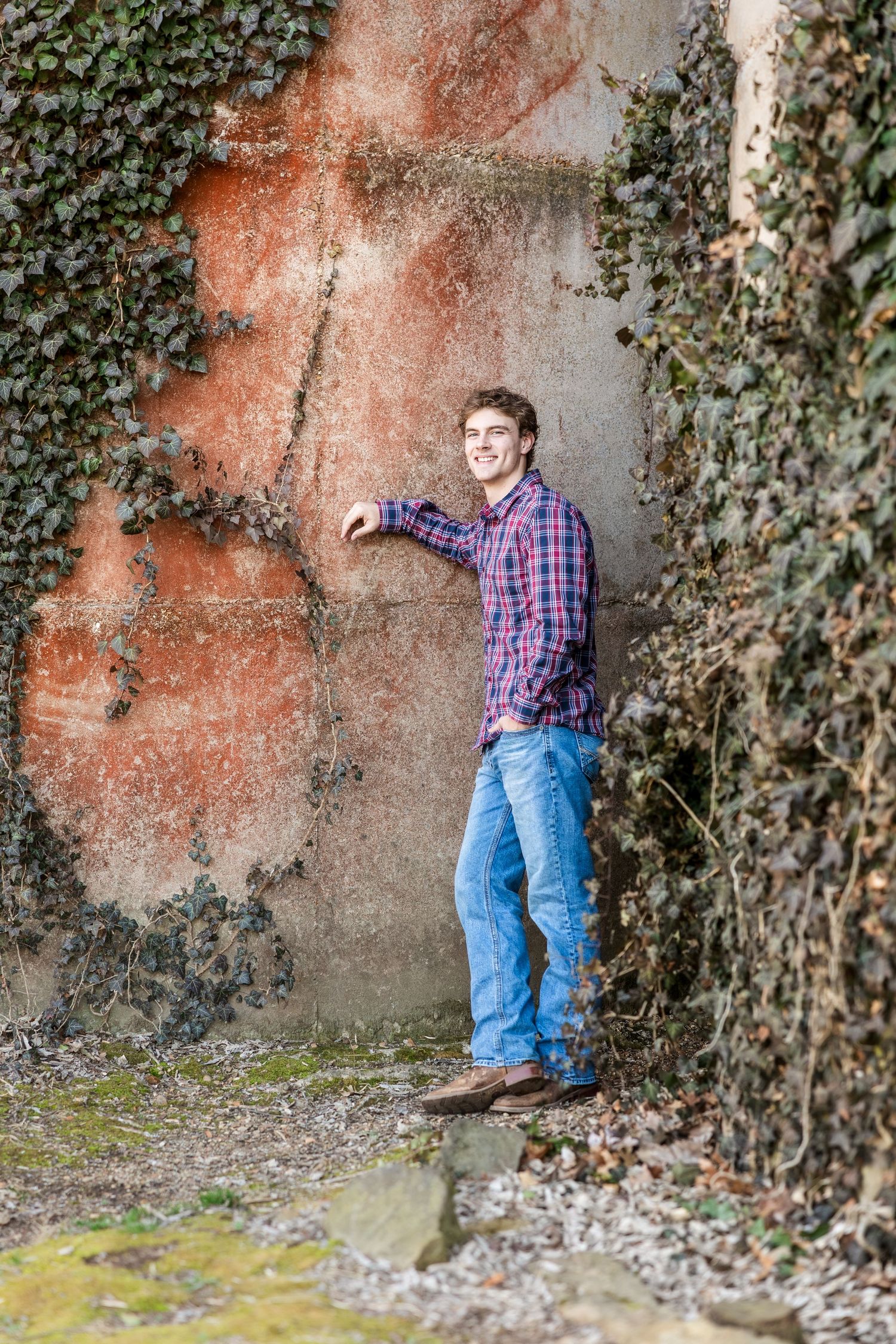 A person in a plaid shirt and jeans stands against a wall covered in ivy and orange staining.