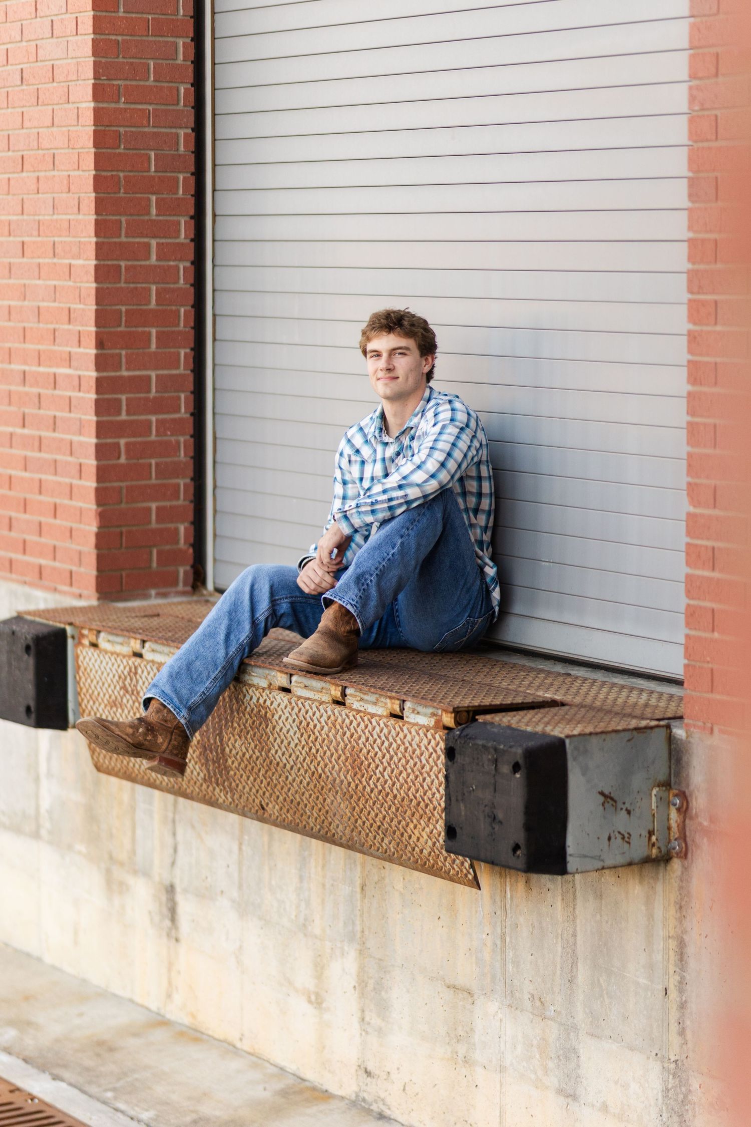 A person in a plaid shirt and jeans sits on a loading dock ledge against brick and metal walls.