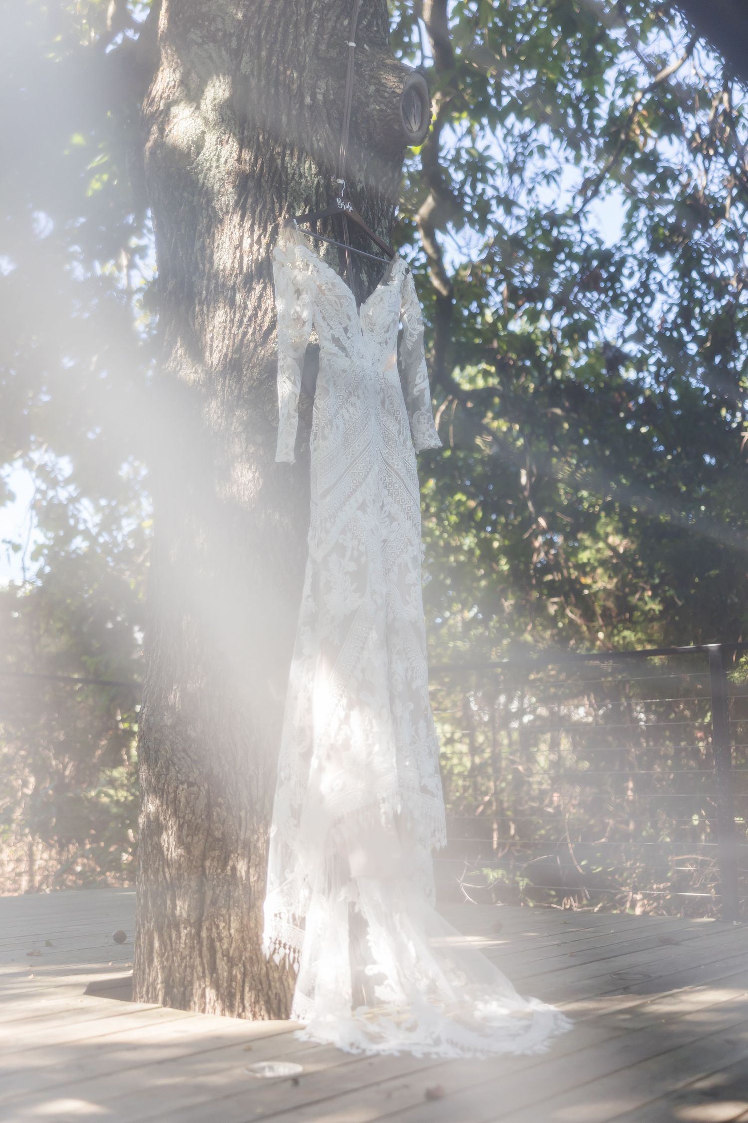 A white wedding dress hangs elegantly from a tree trunk in dappled sunlight creating an ethereal outdoor display.
