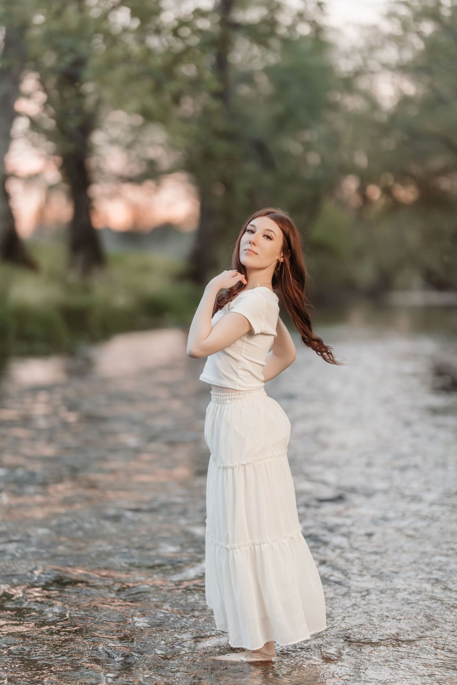Woman in flowing white dress poses gracefully by water during sunset.