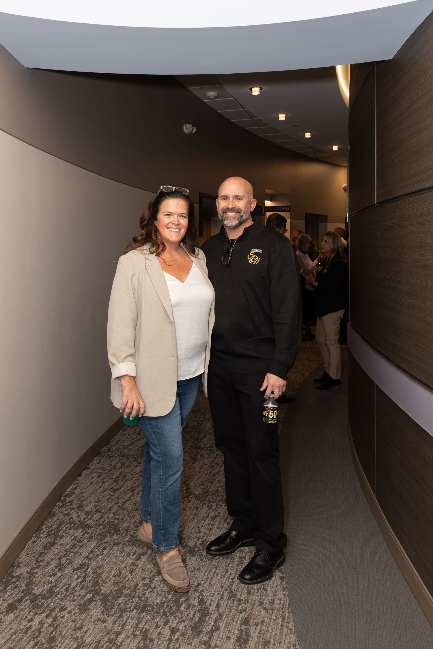 Two people standing together in a modern office hallway with carpeted floors.
