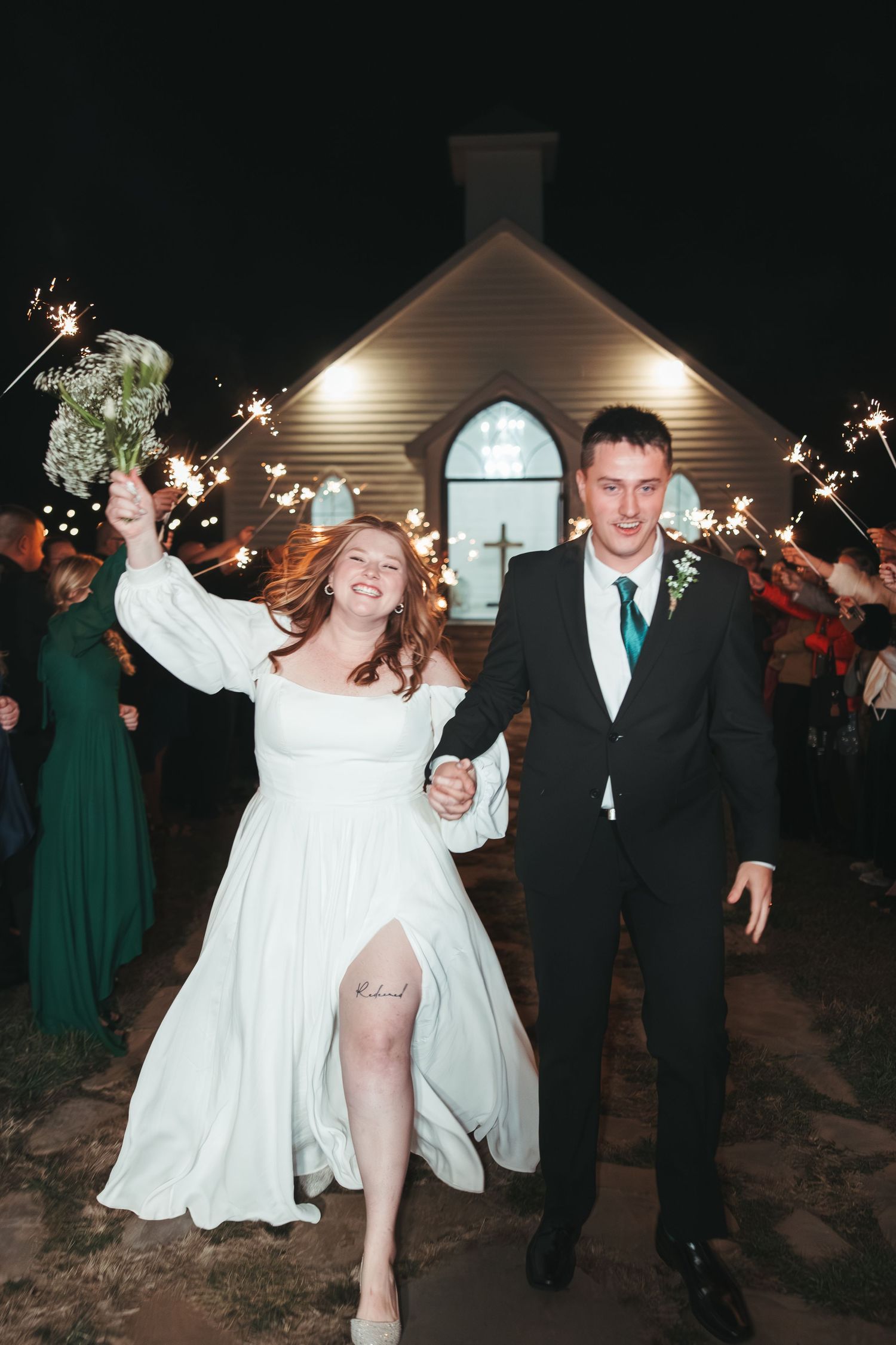 Newlyweds celebrate outside chapel at night surrounded by sparklers.
