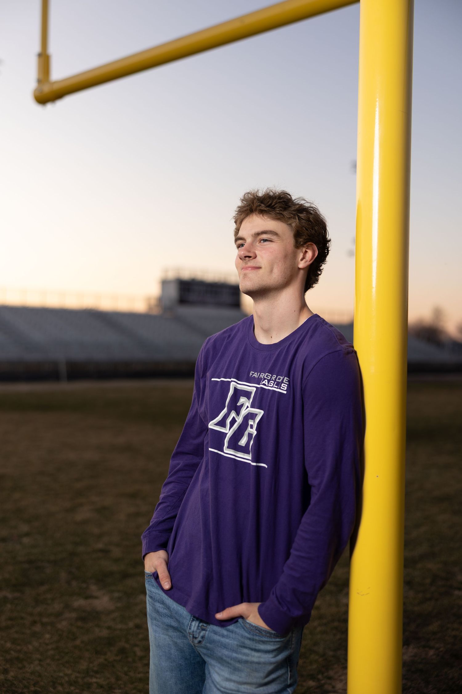 High school student in purple long sleeve shirt leans against yellow football goalpost during sunset.