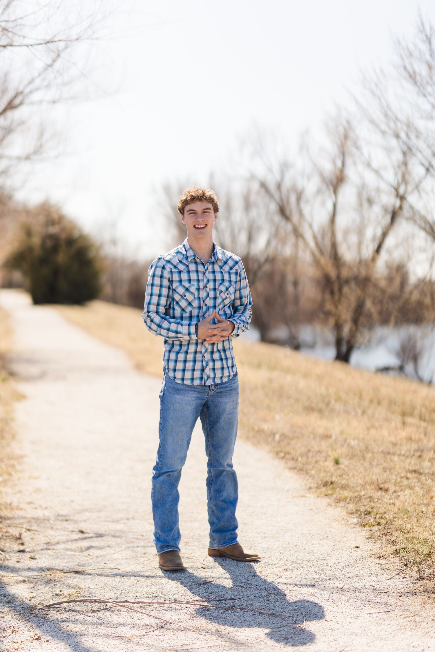 Person standing on dirt path wearing blue plaid shirt and jeans with winter trees in background.