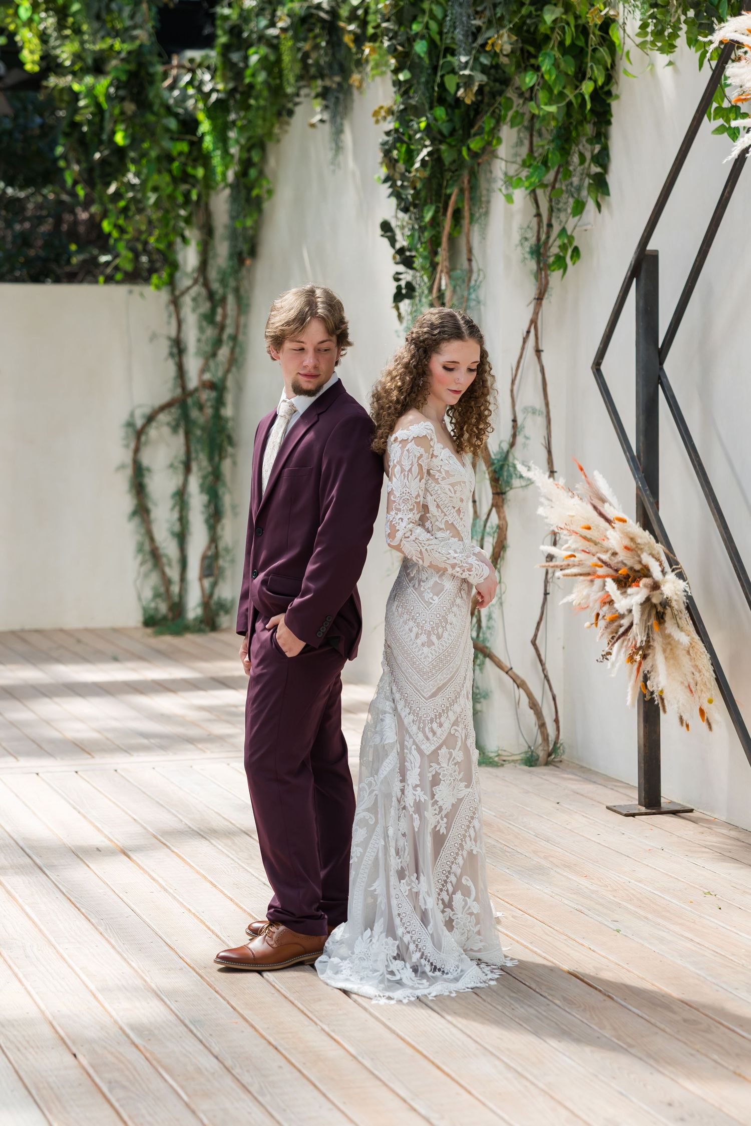 Bride and groom stand back-to-back in lace gown and burgundy suit near green ivy-covered wall.