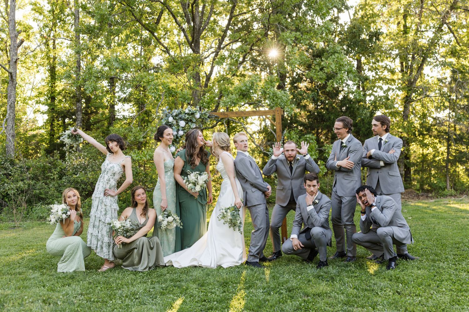 Wedding party celebrates outdoors on lush green lawn with bridesmaids in sage gowns and groomsmen in gray suits under sunlit trees.
