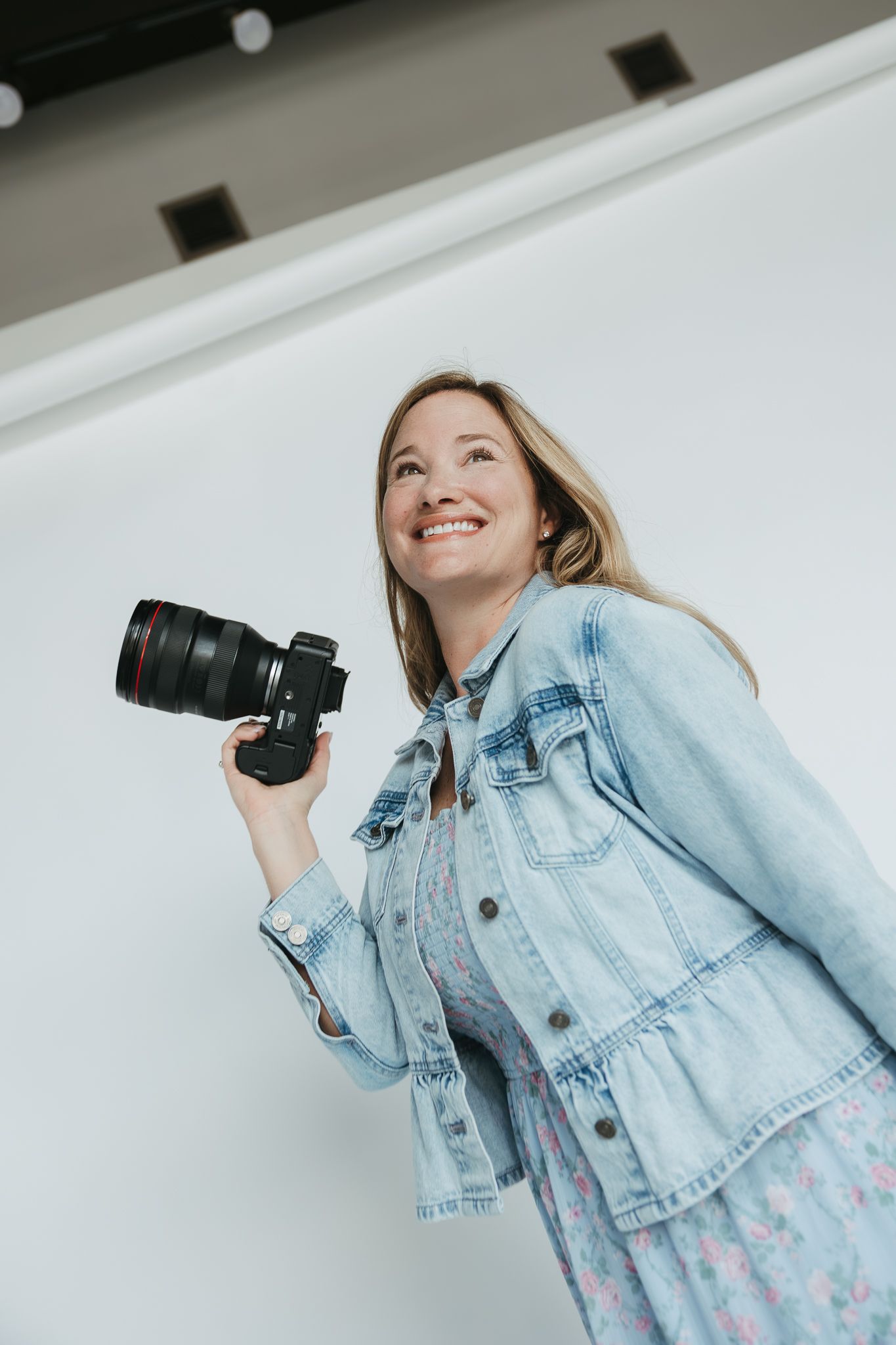 Photographer in a denim jacket holding a professional camera and smiling while looking up against a white wall.