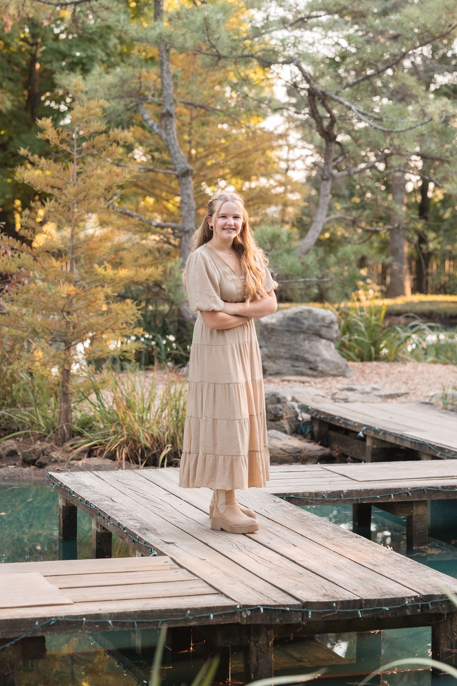 A person in a beige tiered dress stands on a wooden dock over water in an autumn setting.
