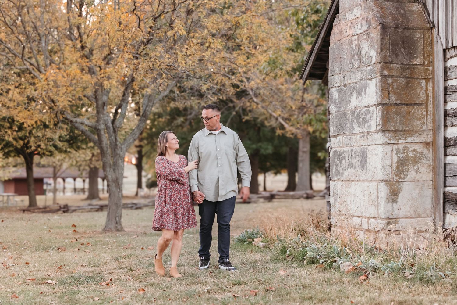 A couple walks together through a rustic autumn setting near an old stone building.
