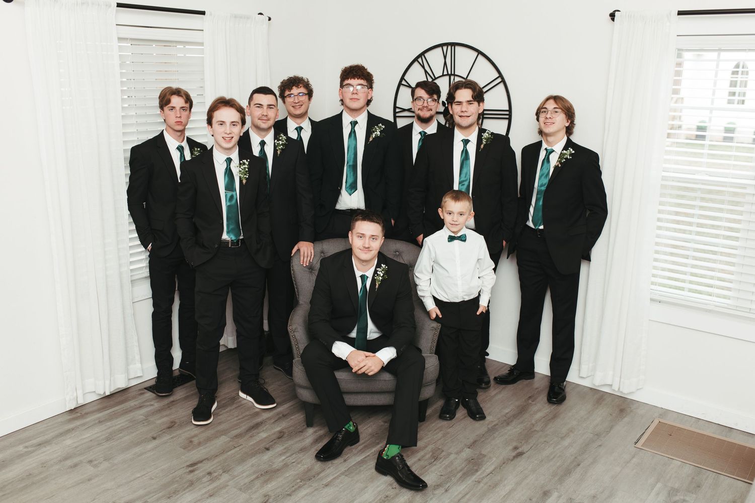 Group in matching black suits and teal ties pose together in bright room with white walls.