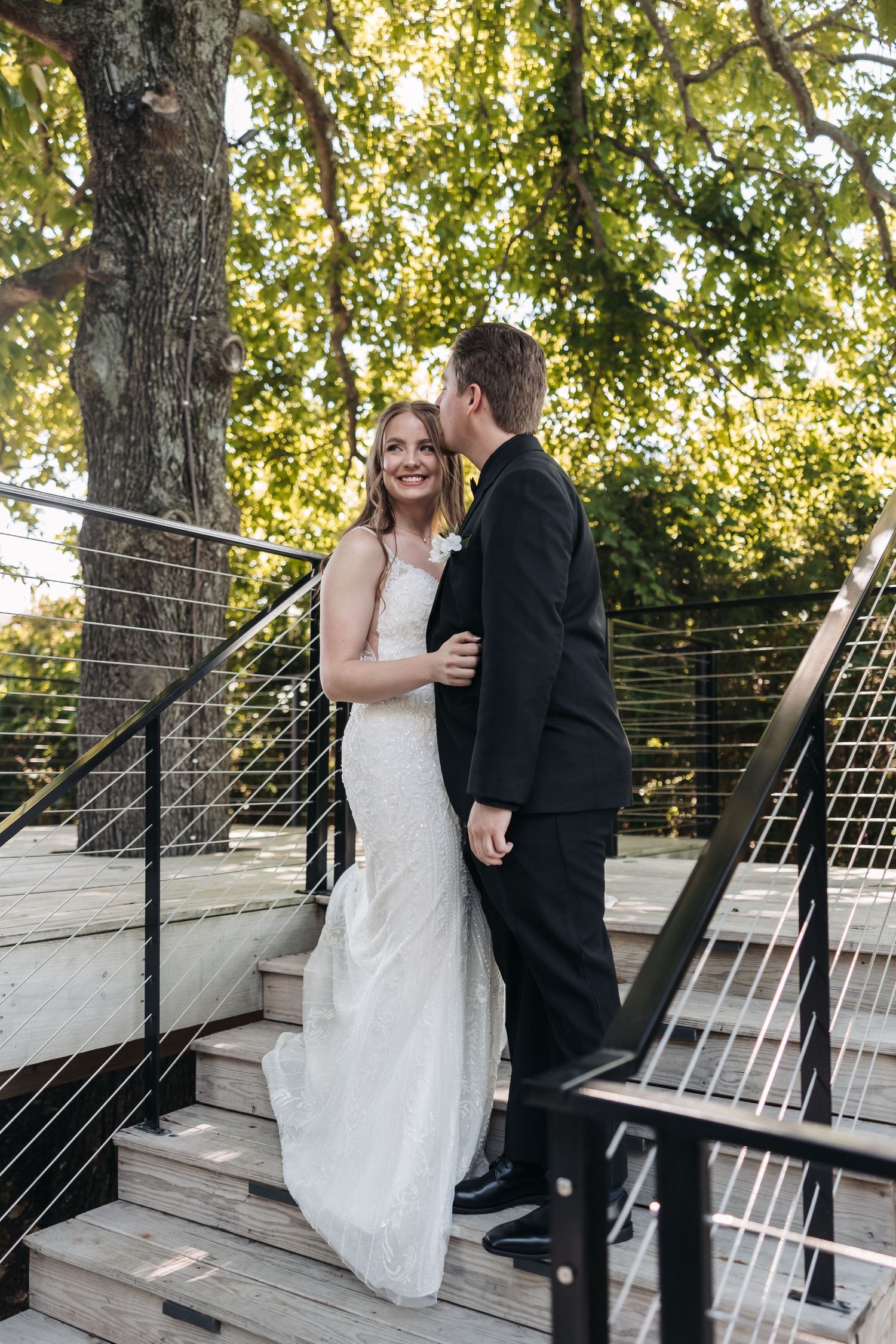 Wedding couple embraces on outdoor wooden deck surrounded by trees.