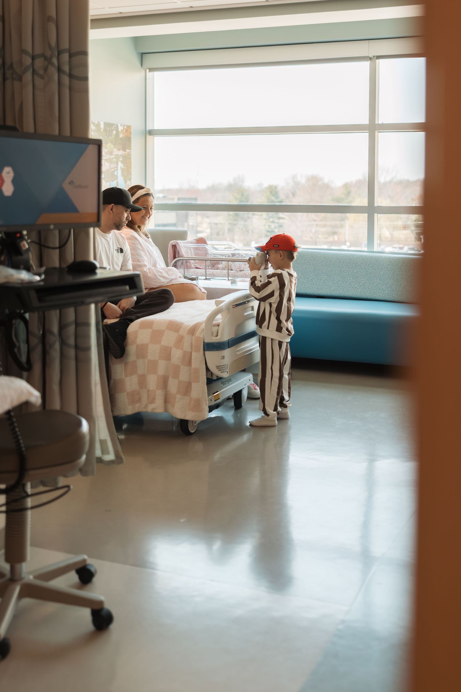 Patient room in hospital with several beds next to windows, medical equipment, and staff attending in background.